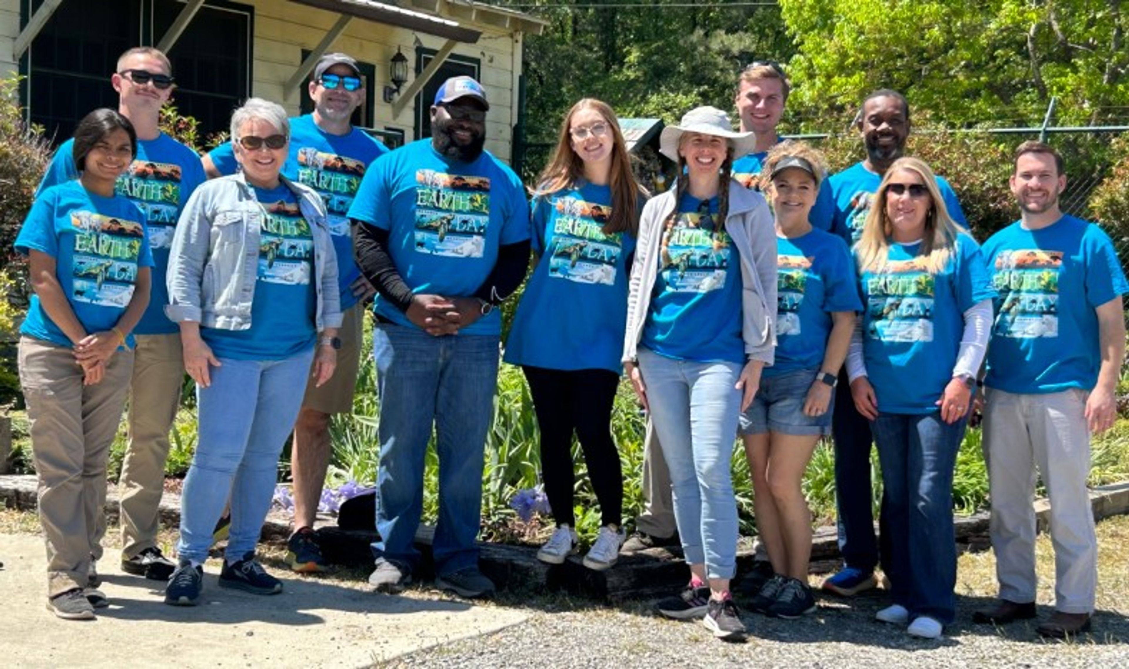 A group of twelve people wearing matching blue T-shirts stands outside in sunlight, smiling at the camera. Behind them is a garden and an old building.