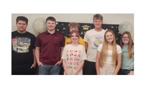 Seven teenagers stand side by side, smiling, in front of a decorated wall with balloons and a graduation cap banner.
