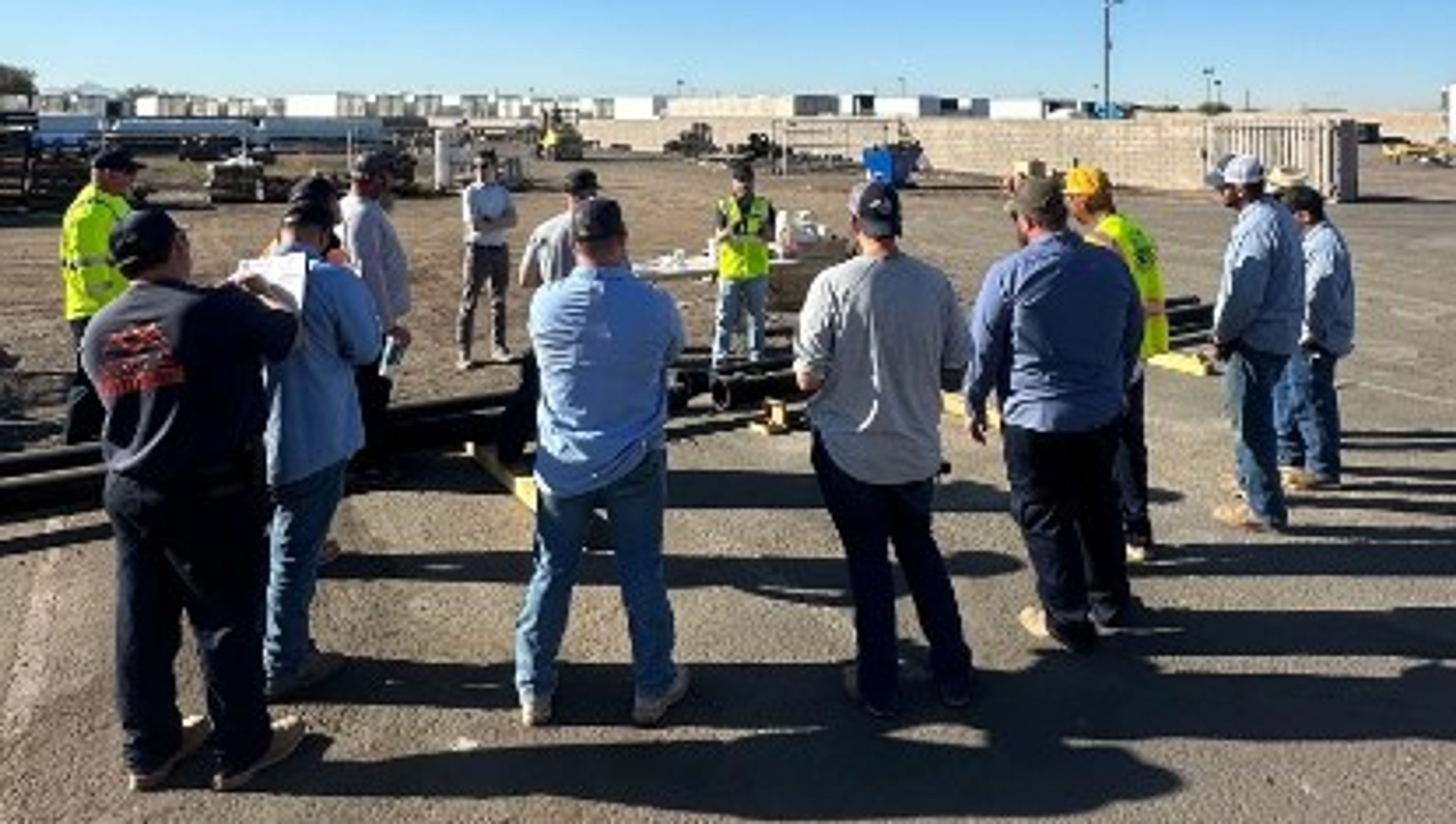 A group of workers in safety gear stand in a circle outdoors on a sunny day, listening to a person speaking in front of them at a construction or industrial site.