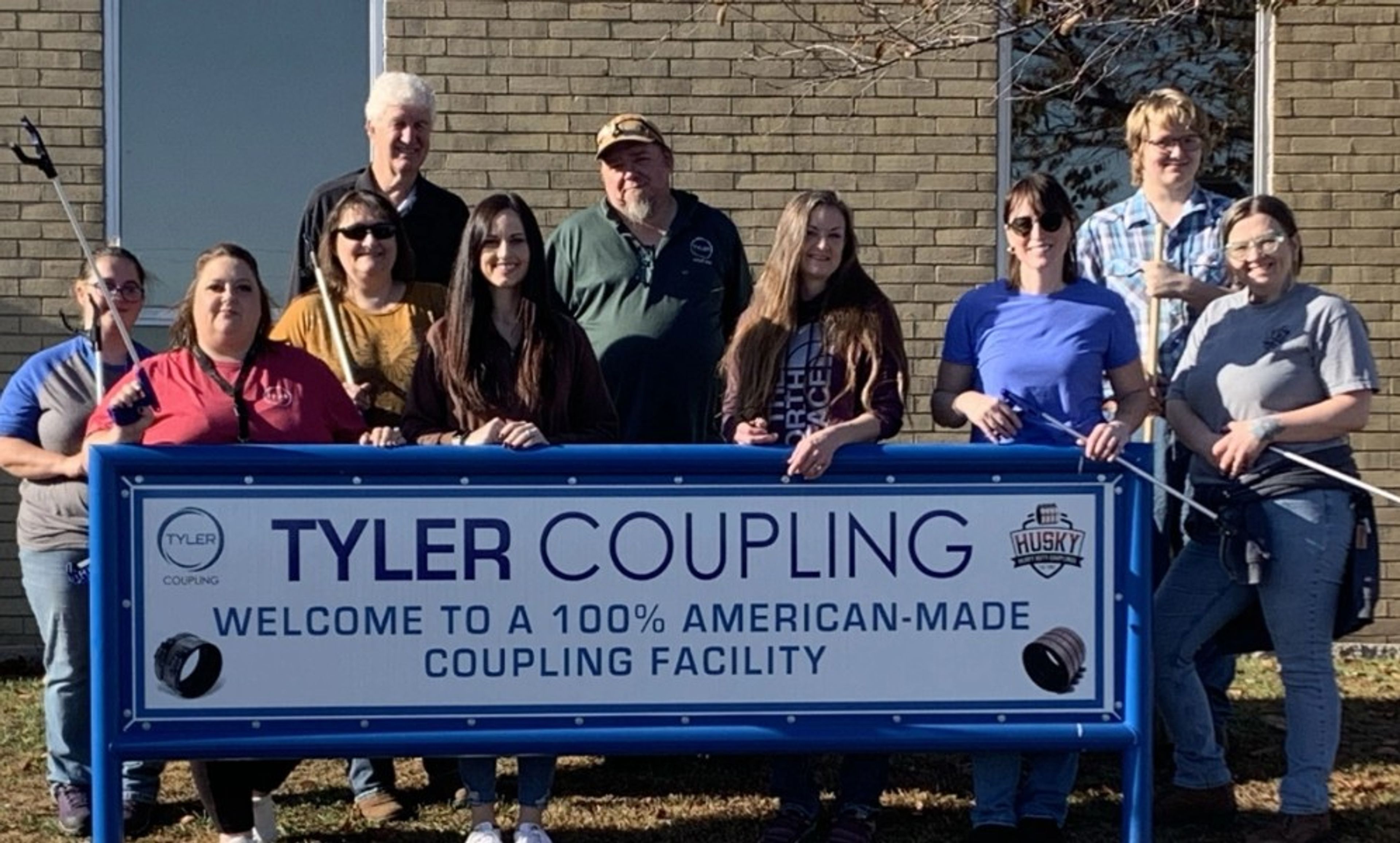 A group of ten people stand behind a blue sign that reads, “Tyler Coupling. Welcome to a 100% American-made coupling facility,” in front of a brick building on a sunny day.