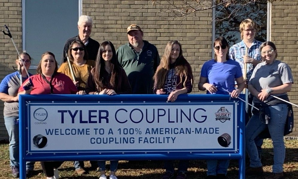 A group of ten people stand behind a blue sign that reads, “Tyler Coupling. Welcome to a 100% American-made coupling facility,” in front of a brick building on a sunny day.