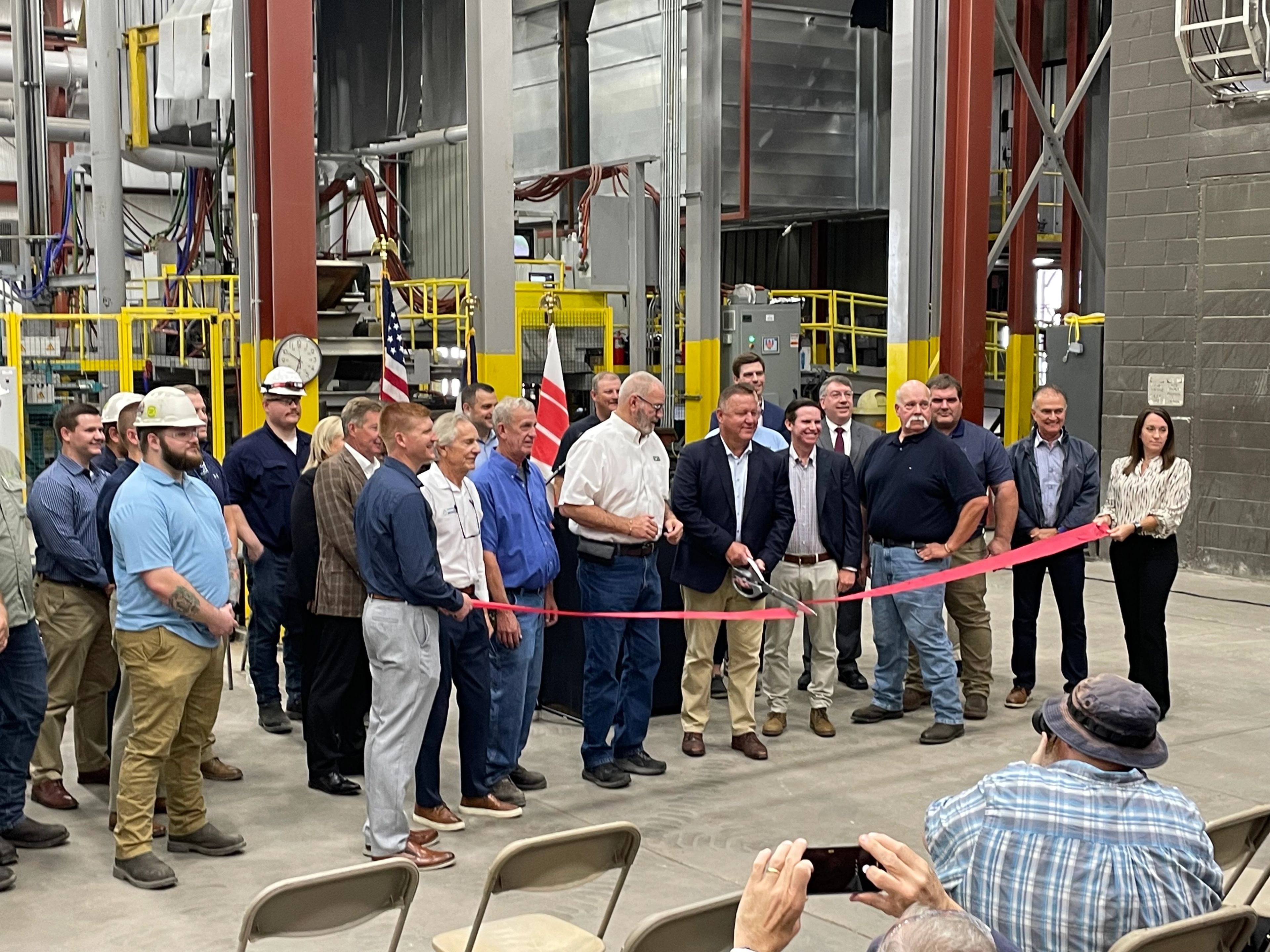 A group of people, some in business attire and others in work uniforms, gather indoors for a ribbon-cutting ceremony at an industrial facility, with an audience seated in front.