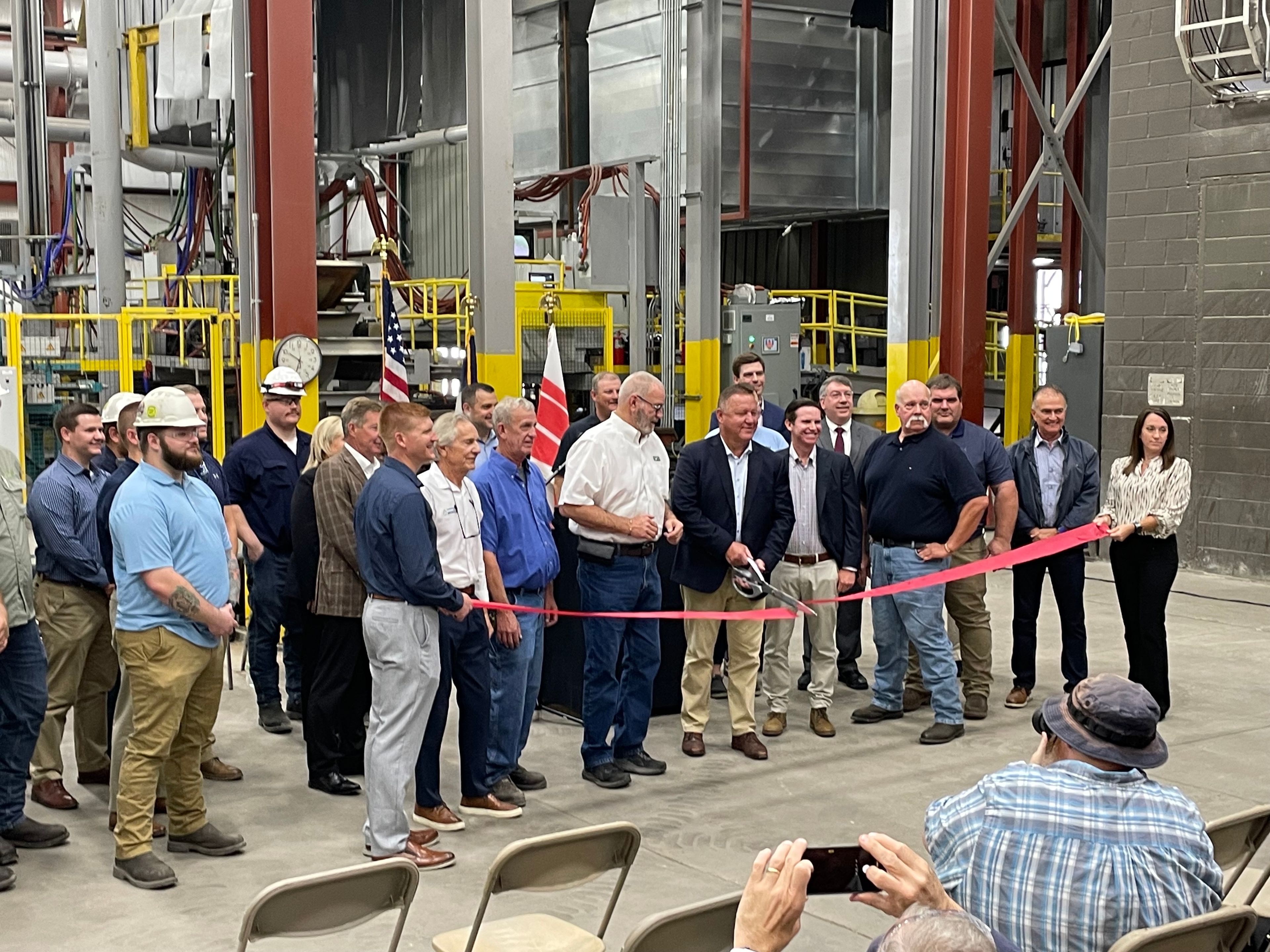 A group of people, some in business attire and others in work uniforms, gather indoors for a ribbon-cutting ceremony at an industrial facility, with an audience seated in front.