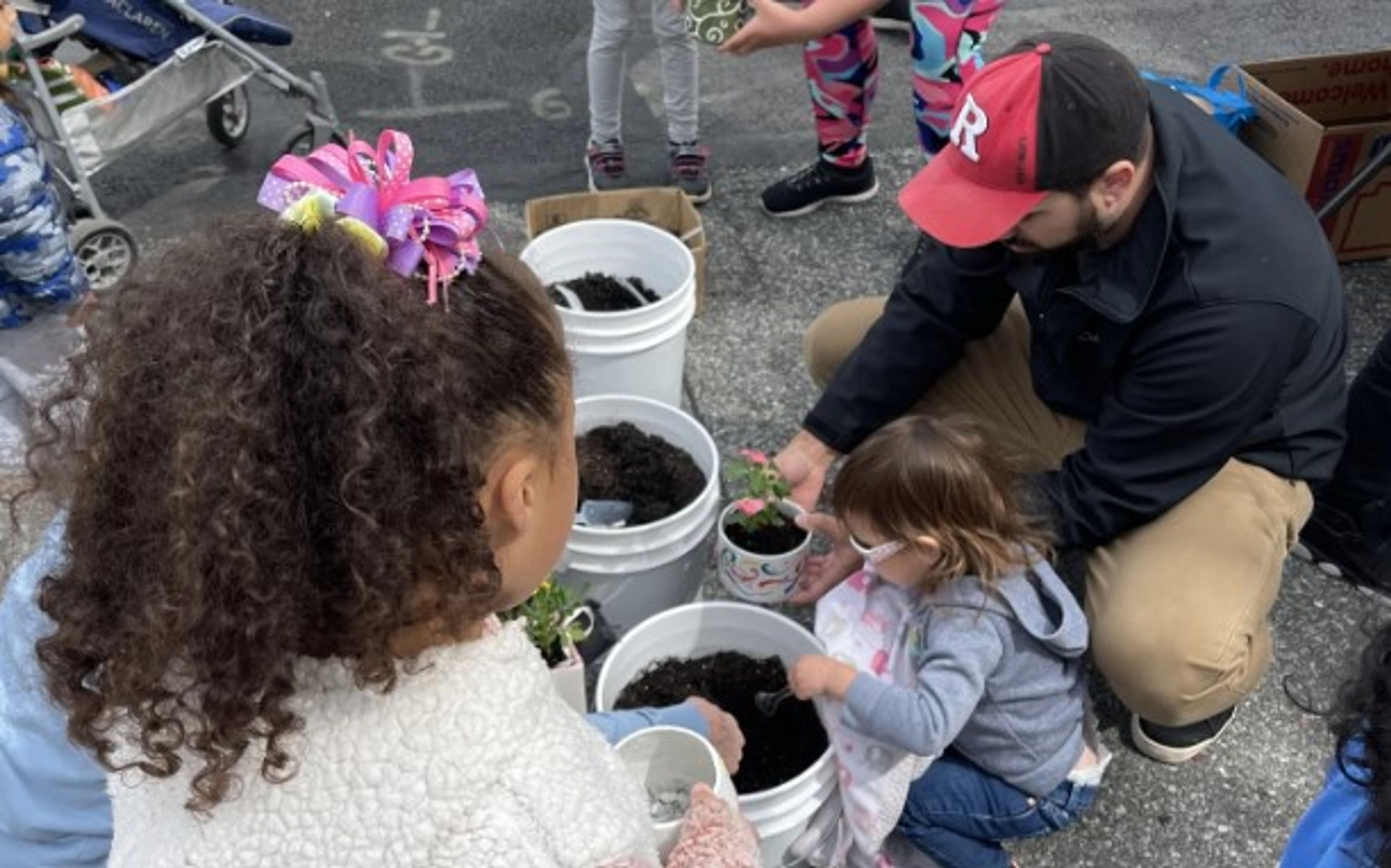 Adults and children gather outdoors around white buckets filled with soil, planting small flowers together. A man in a red cap helps a child while others watch and participate. The activity takes place on pavement.