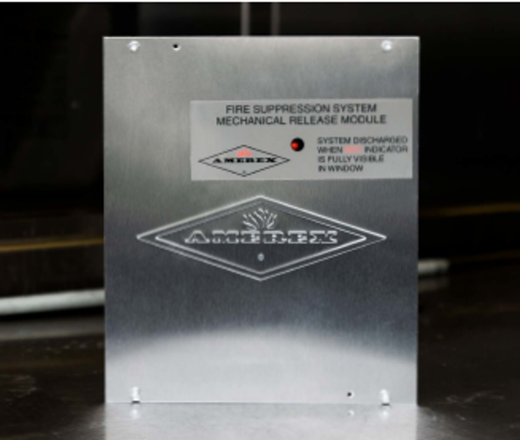 A metal Amerex fire suppression system mechanical release module with a label indicating system status and a visible indicator window.