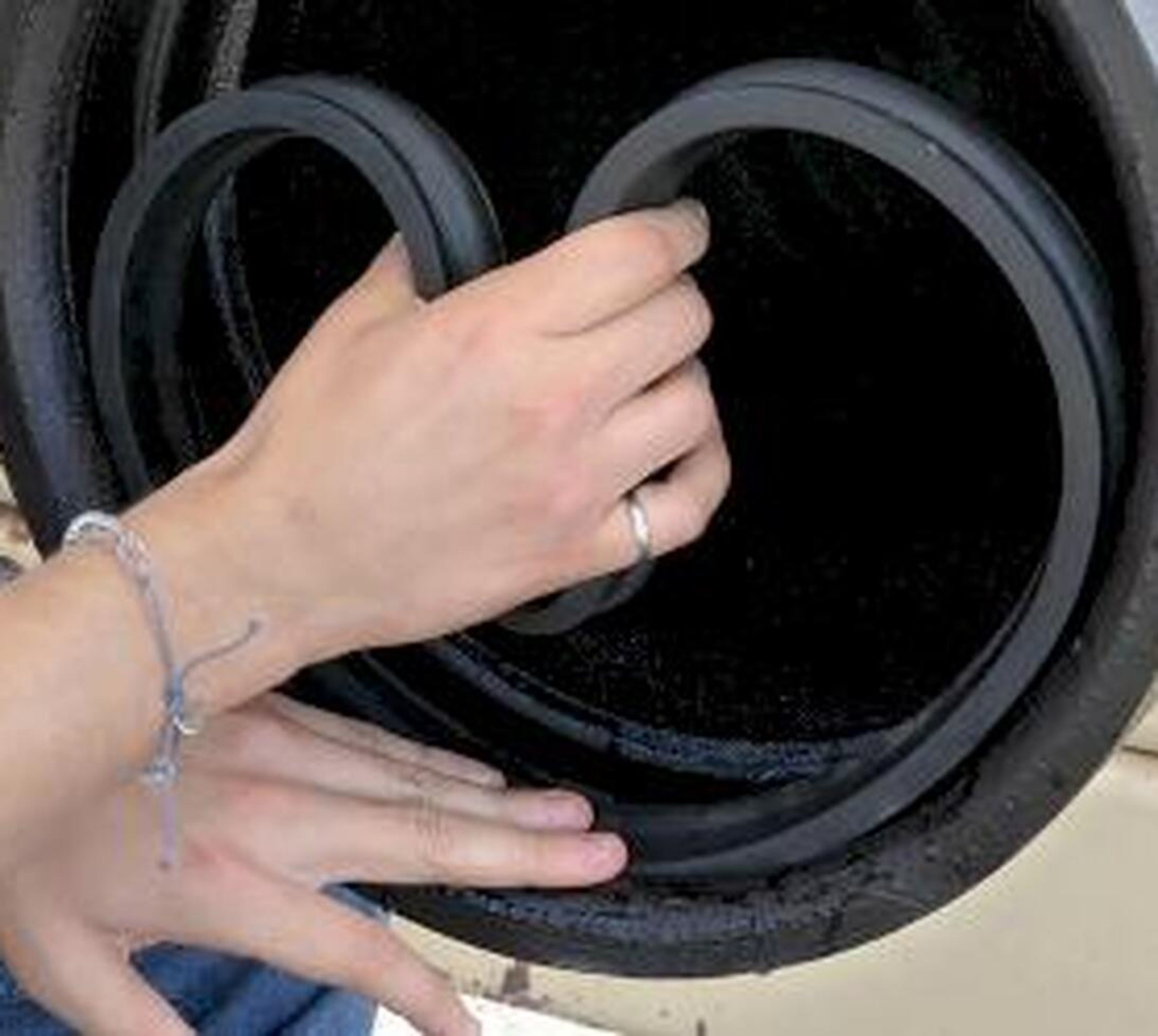 Two hands fitting a circular rubber seal onto the edge of a black cylindrical object. The person wears a silver ring and a thin bracelet on their left wrist.