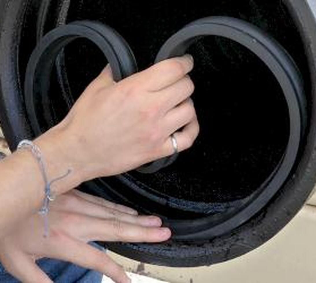 Two hands fitting a circular rubber seal onto the edge of a black cylindrical object. The person wears a silver ring and a thin bracelet on their left wrist.