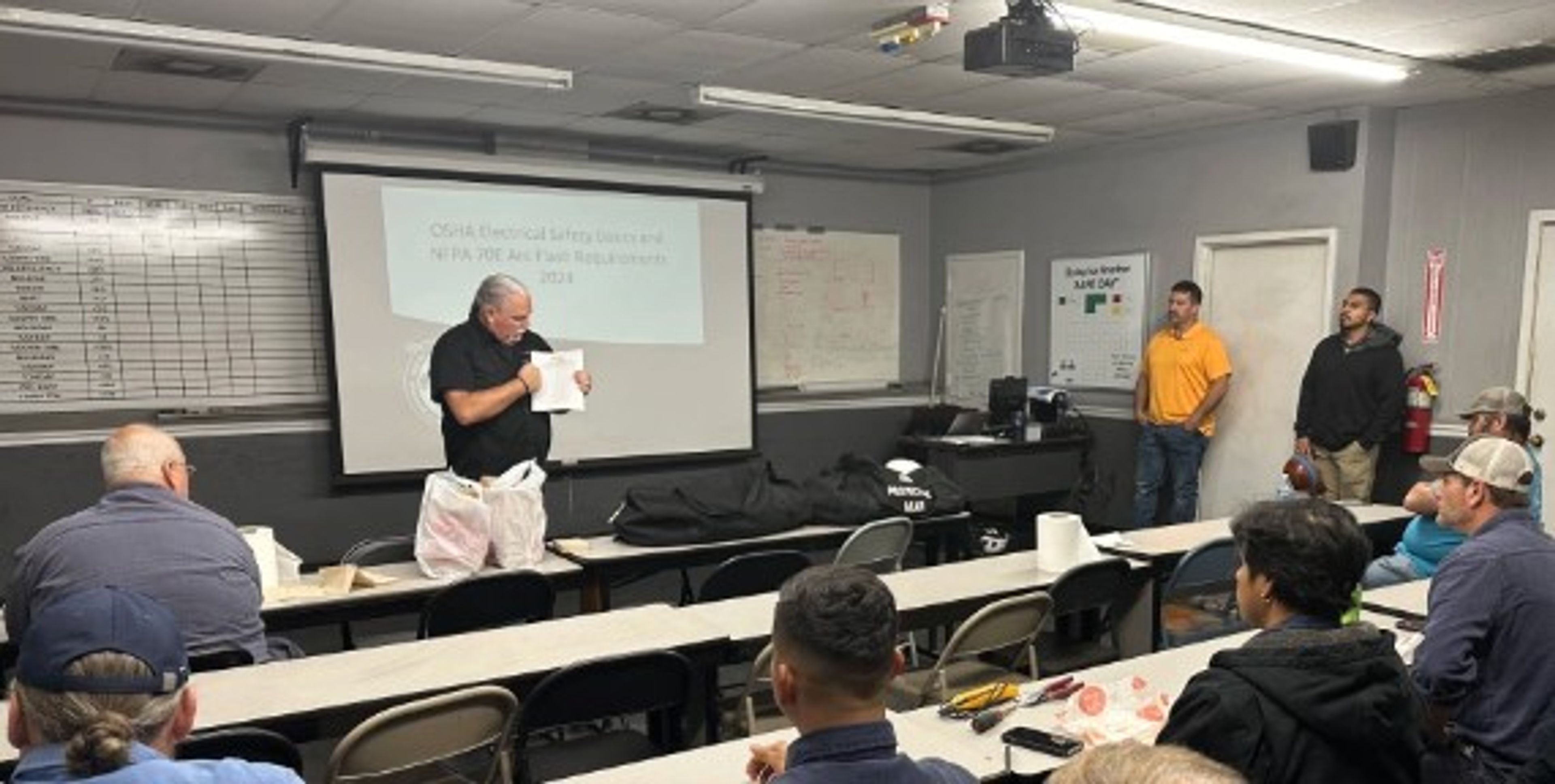 A man presents to a group in a classroom with a projector displaying a slide. Several people are seated at tables, listening. Charts and notes are visible on the walls.