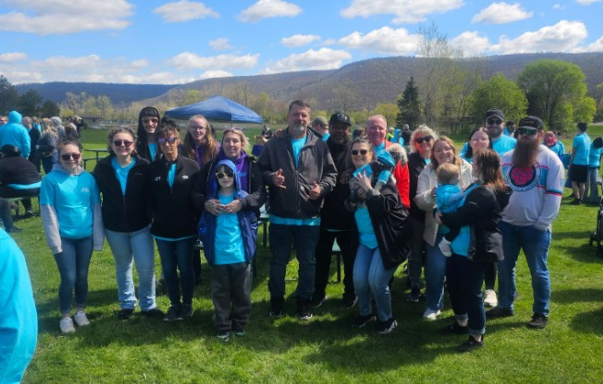 A group of people, many wearing teal shirts, pose together outdoors on a sunny day with mountains and blue sky in the background.