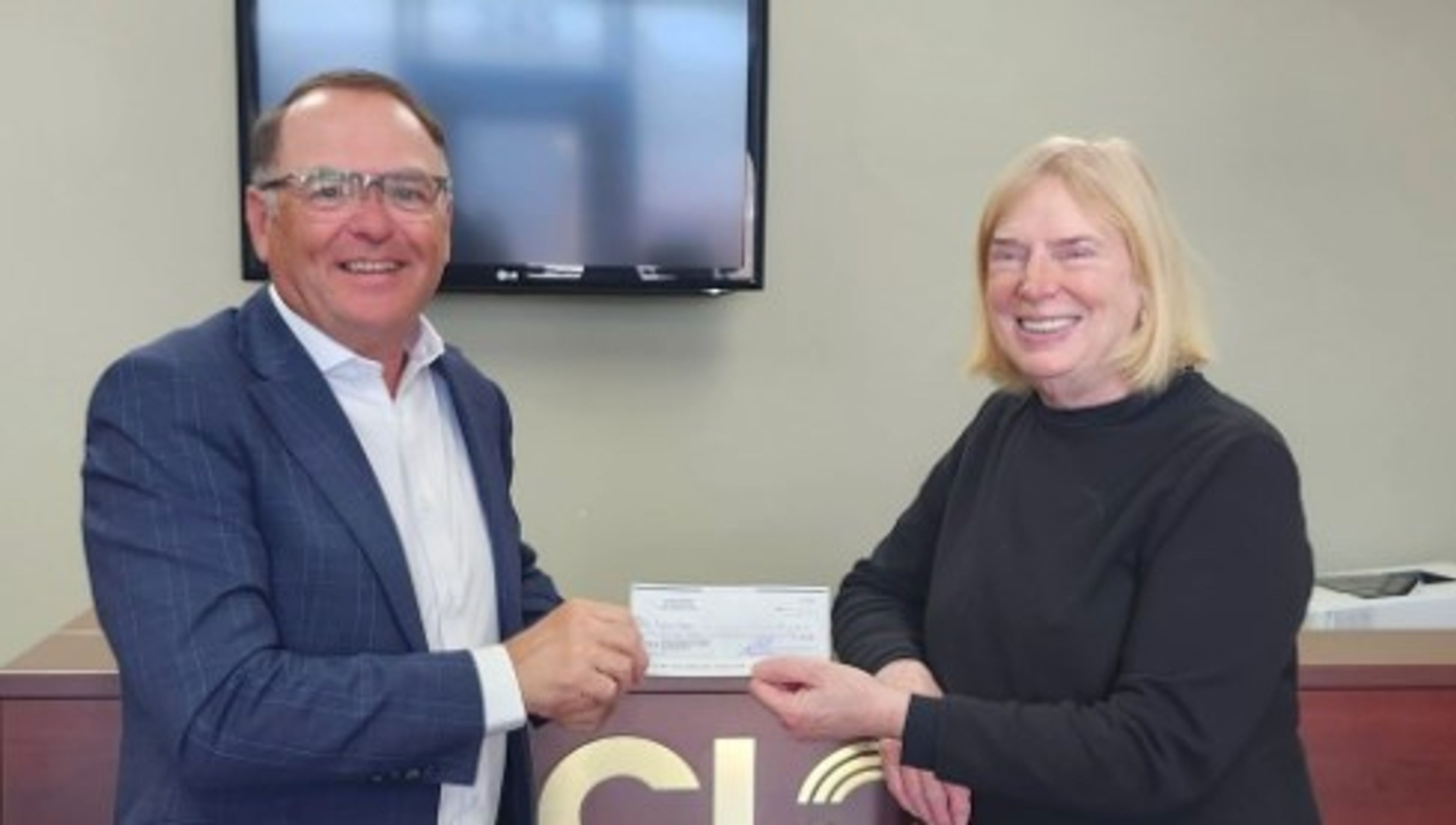 Two people stand indoors, smiling and holding a check together in front of a desk with the letters “CL” visible. A television screen is mounted on the wall behind them.