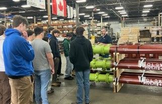 A group of students tours a warehouse, listening to a guide explain large industrial pipes stacked nearby. The warehouse has shelves, boxes, and a Canadian flag visible.