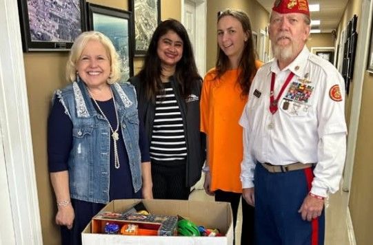 Four adults stand smiling in a hallway behind a box of donated goods. One man wears a military uniform with medals, while the others wear casual clothing. Framed photos hang on the wall behind them.