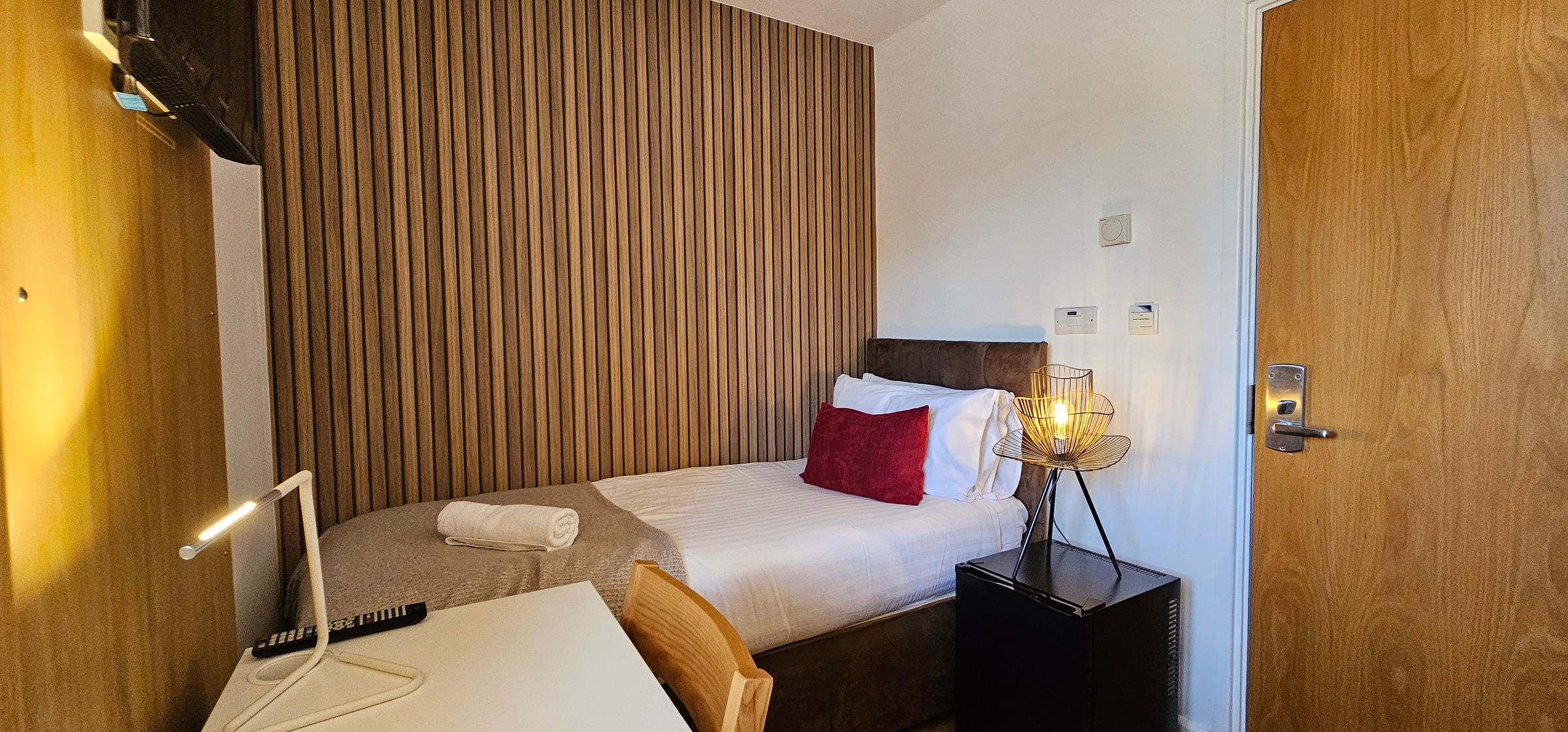Made up single bed with a gold wire lamp on a black fridge cabinet