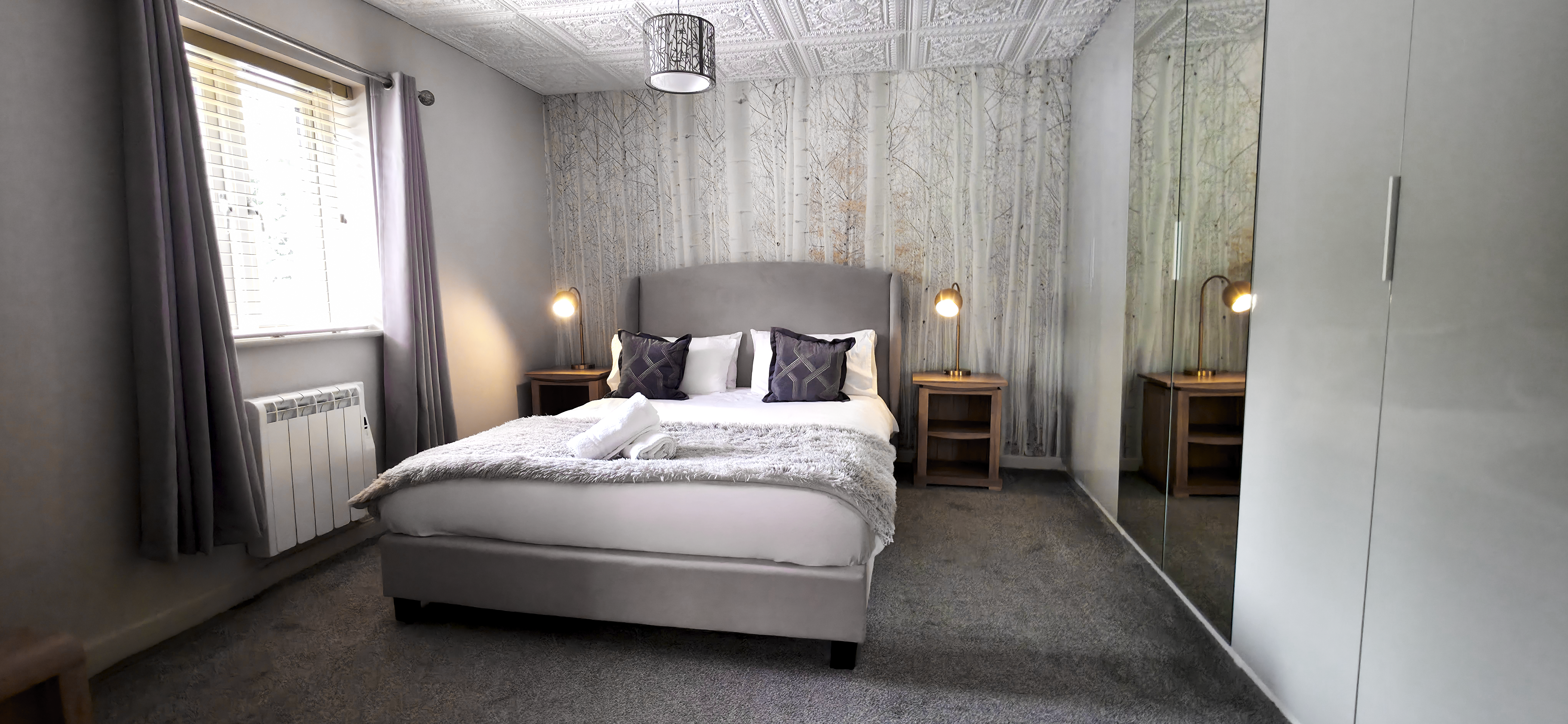 King size bed with upholstered headboard against birch tree wallpaper flanked by brass bedside lamps & walled mirrored wardrobe