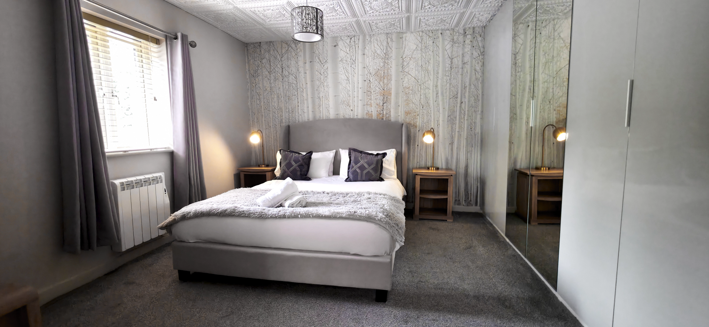 King size bed with upholstered headboard against birch tree wallpaper flanked by brass bedside lamps & walled mirrored wardrobe