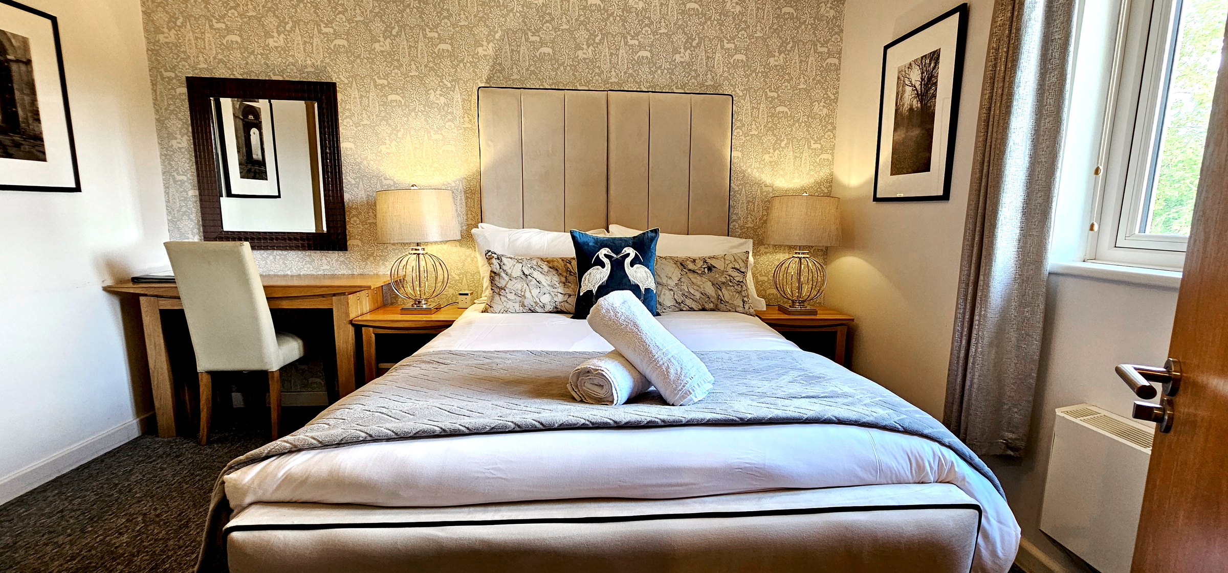 Double bed with panelled headboard between two lamps under patterned wallpaper with framed prints