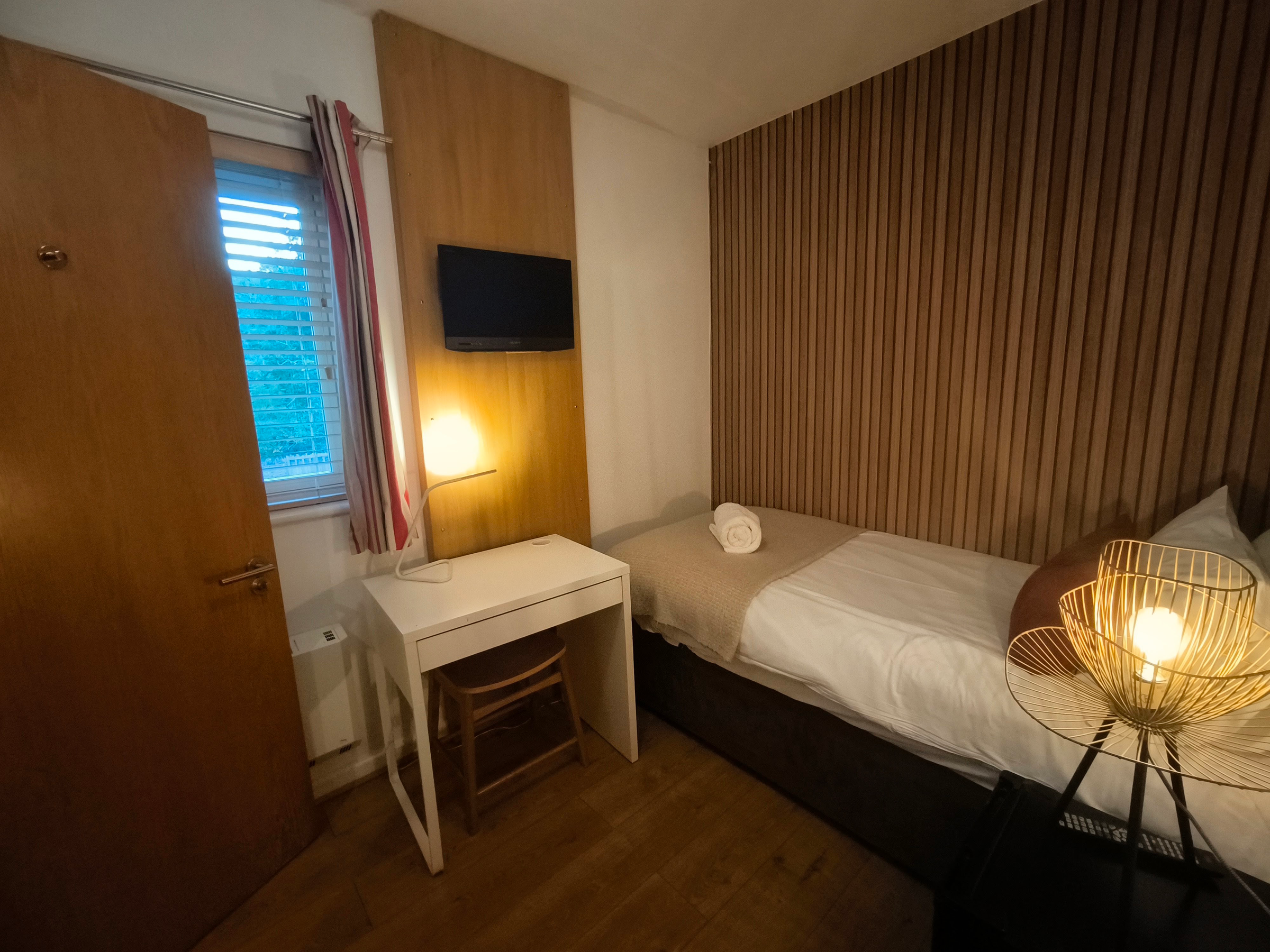 Single room with white desk chair & window with venetian blinds & TV