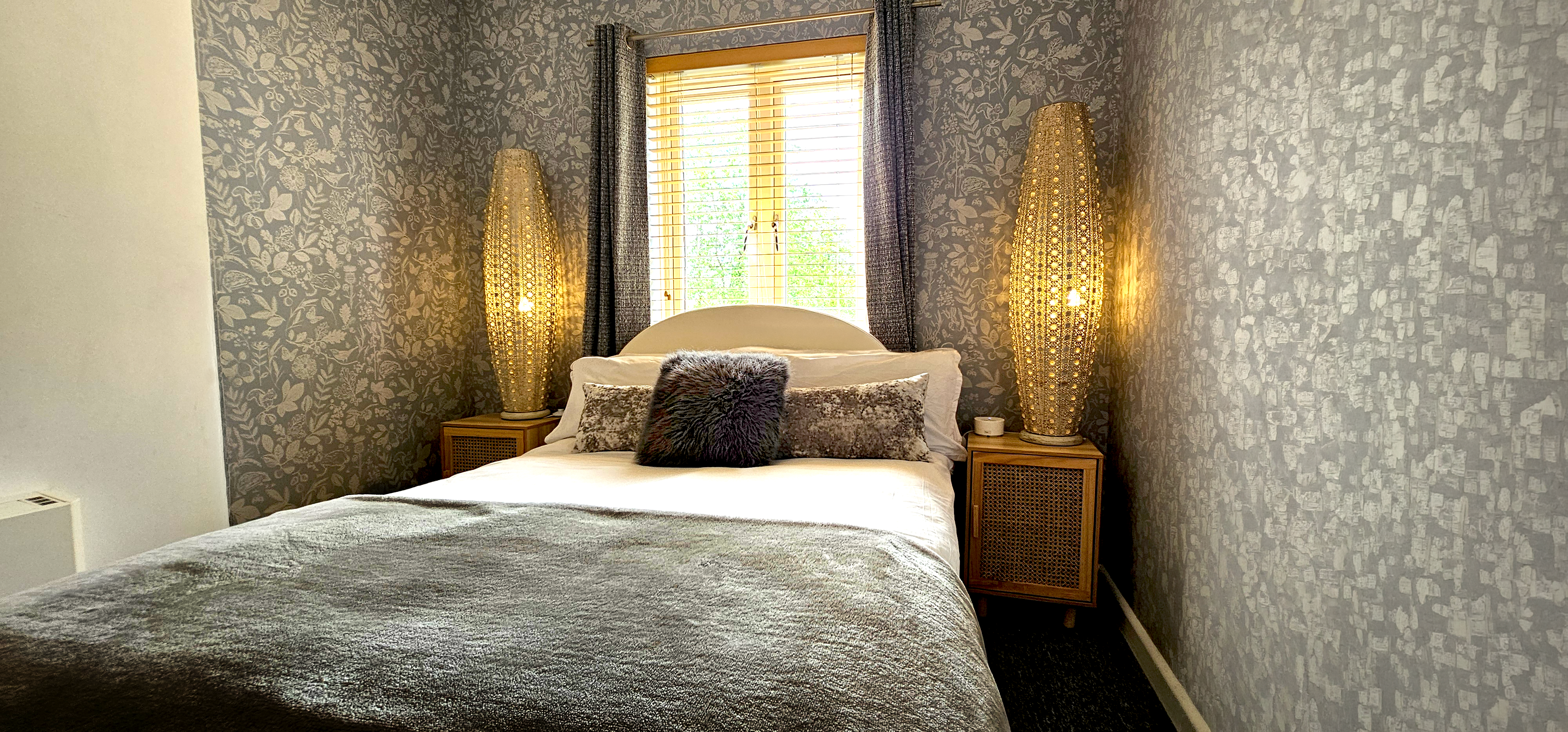 Double bed with grey throw between two woven floor lamps under a shuttered window