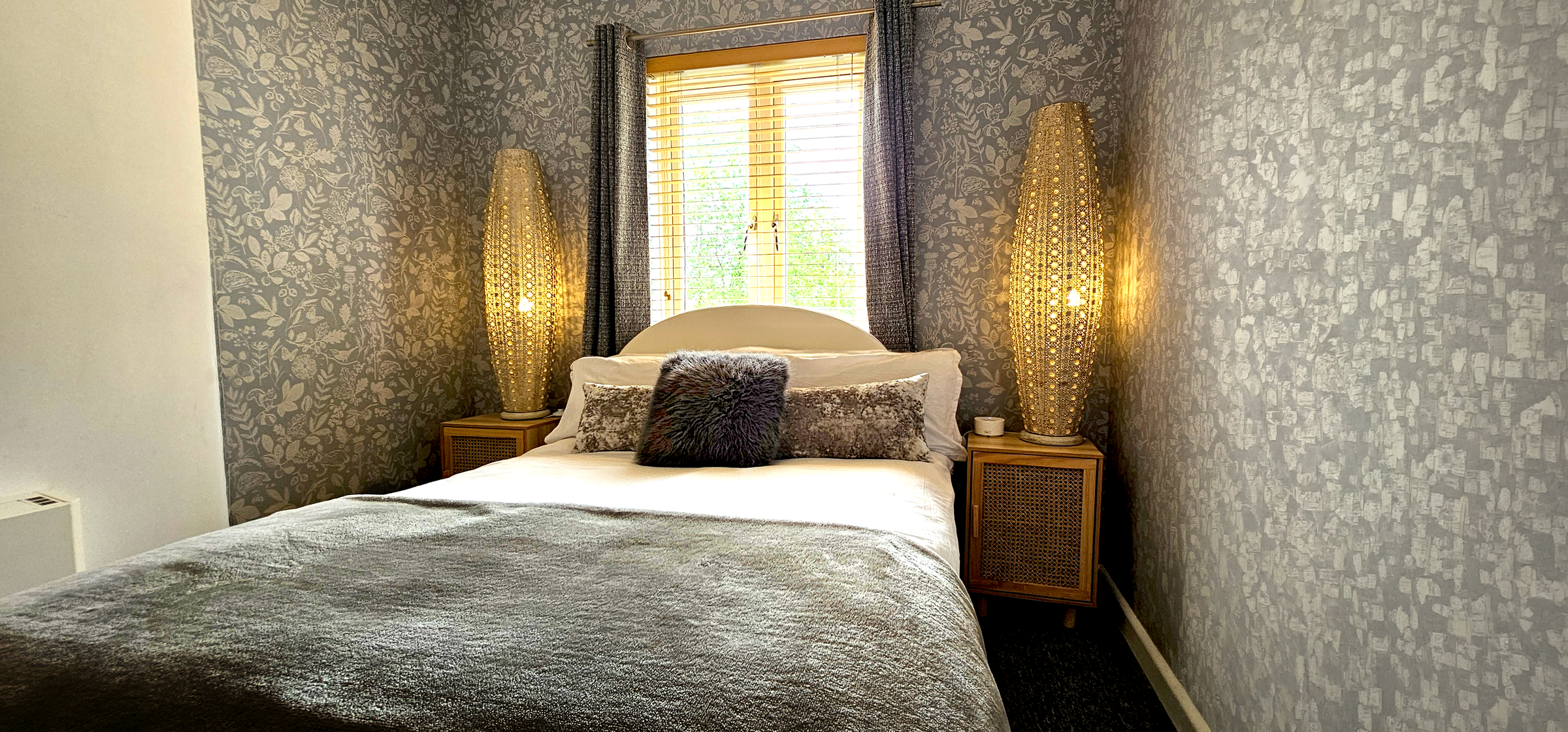 Double bed with grey throw between two woven floor lamps under a shuttered window