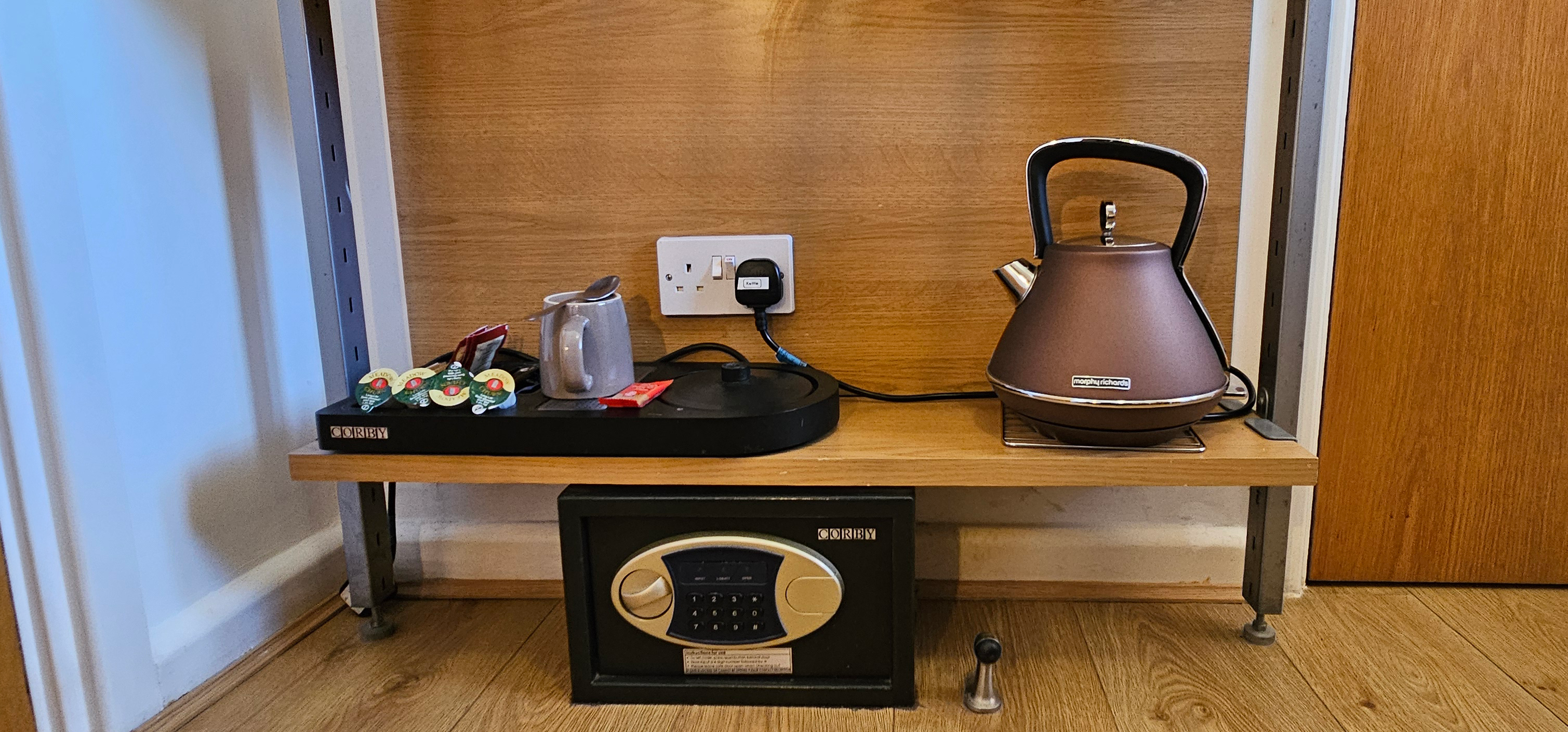 Kettle, cups, tea & coffee sachets on a shelf above a small secure safe & clothes hangers above