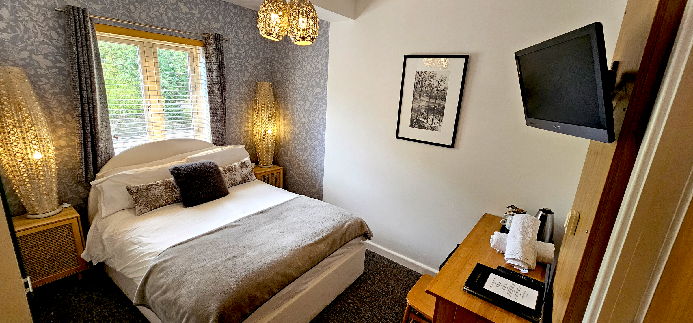 Double bed with grey throw between two woven lamps, digital TV & Desk