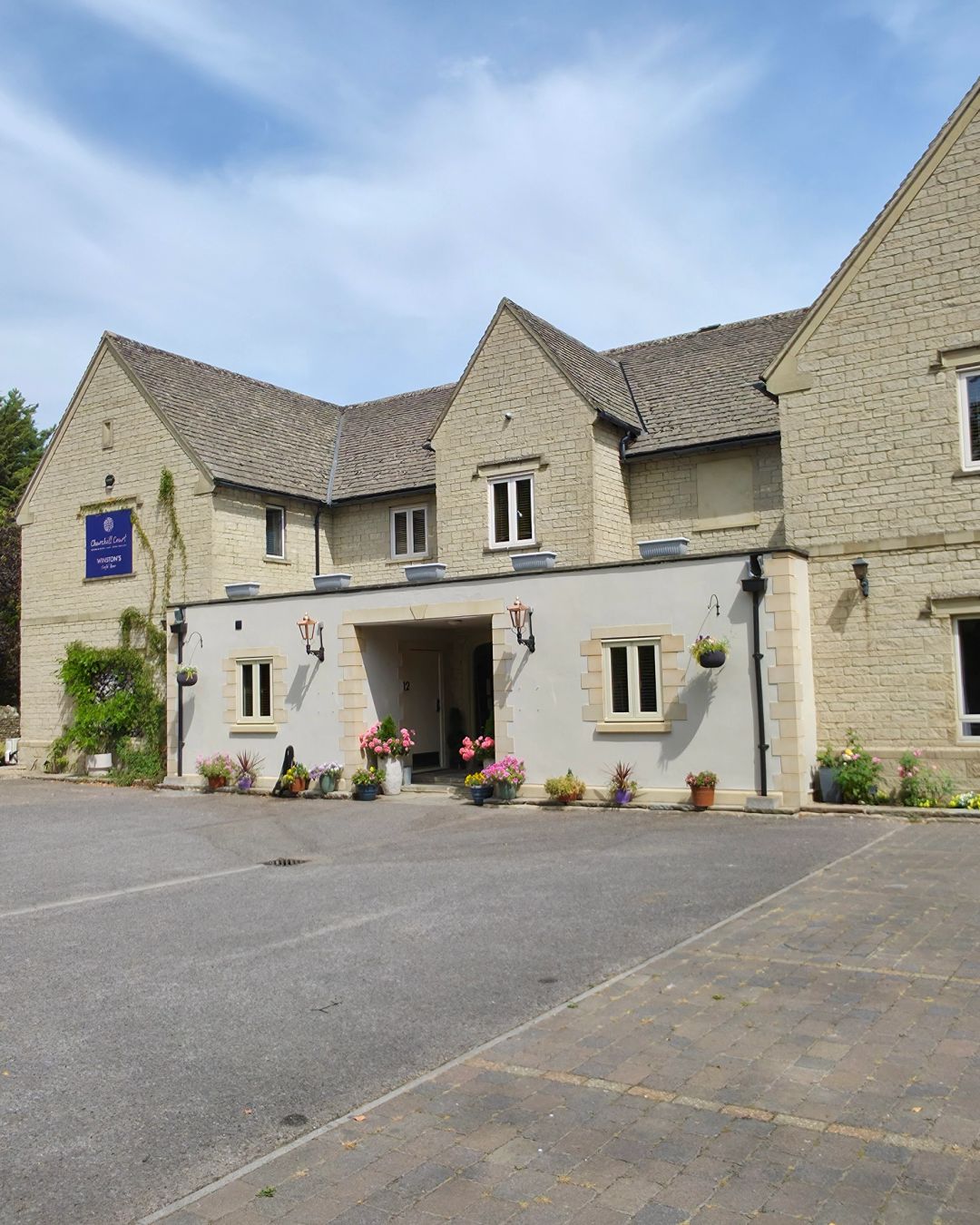 Stone building with covered entrance and planters beside block paved parking area