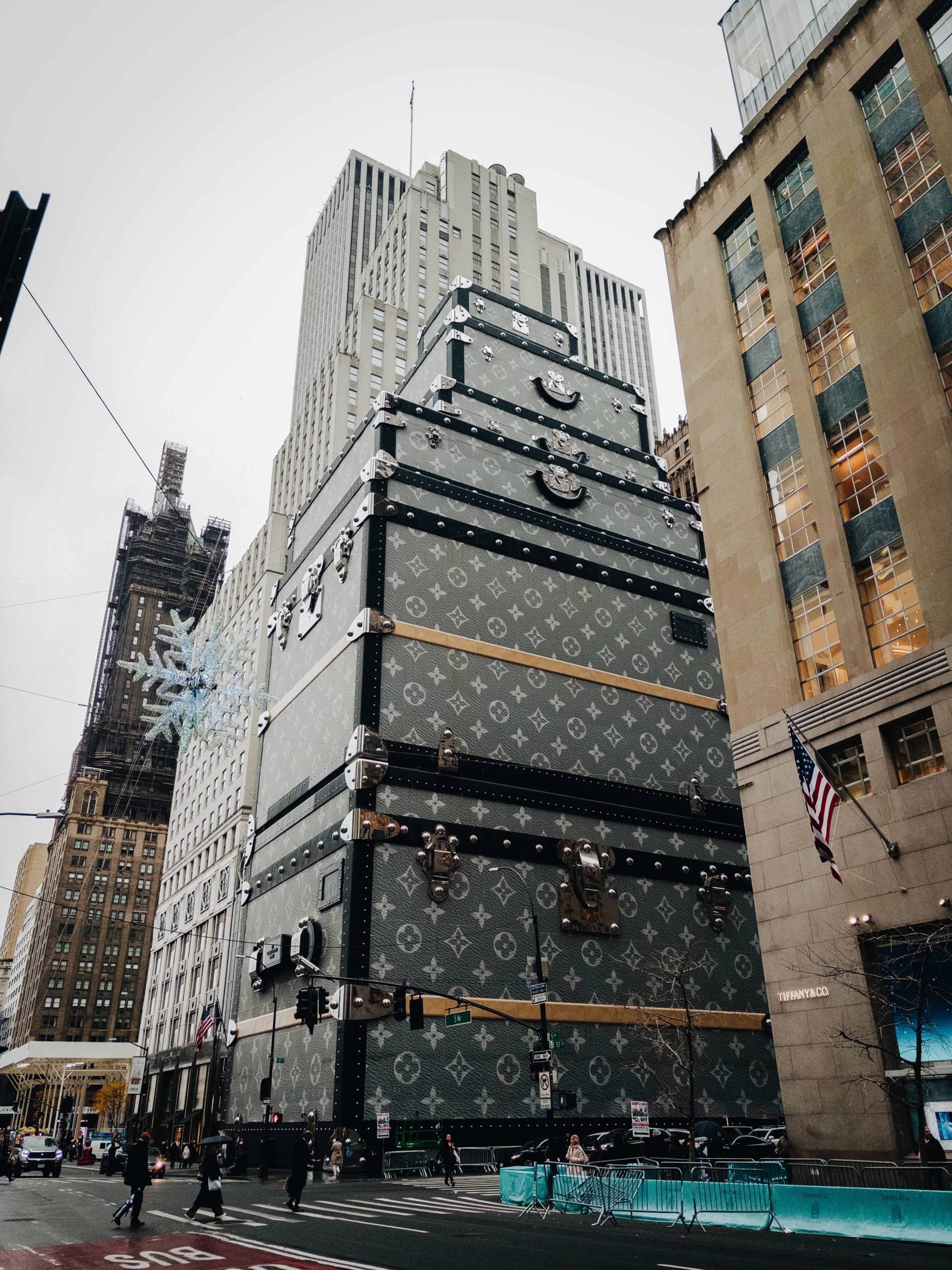 City skyscraper wrapped to resemble a giant monogrammed travel trunk, with pedestrians crossing the street below