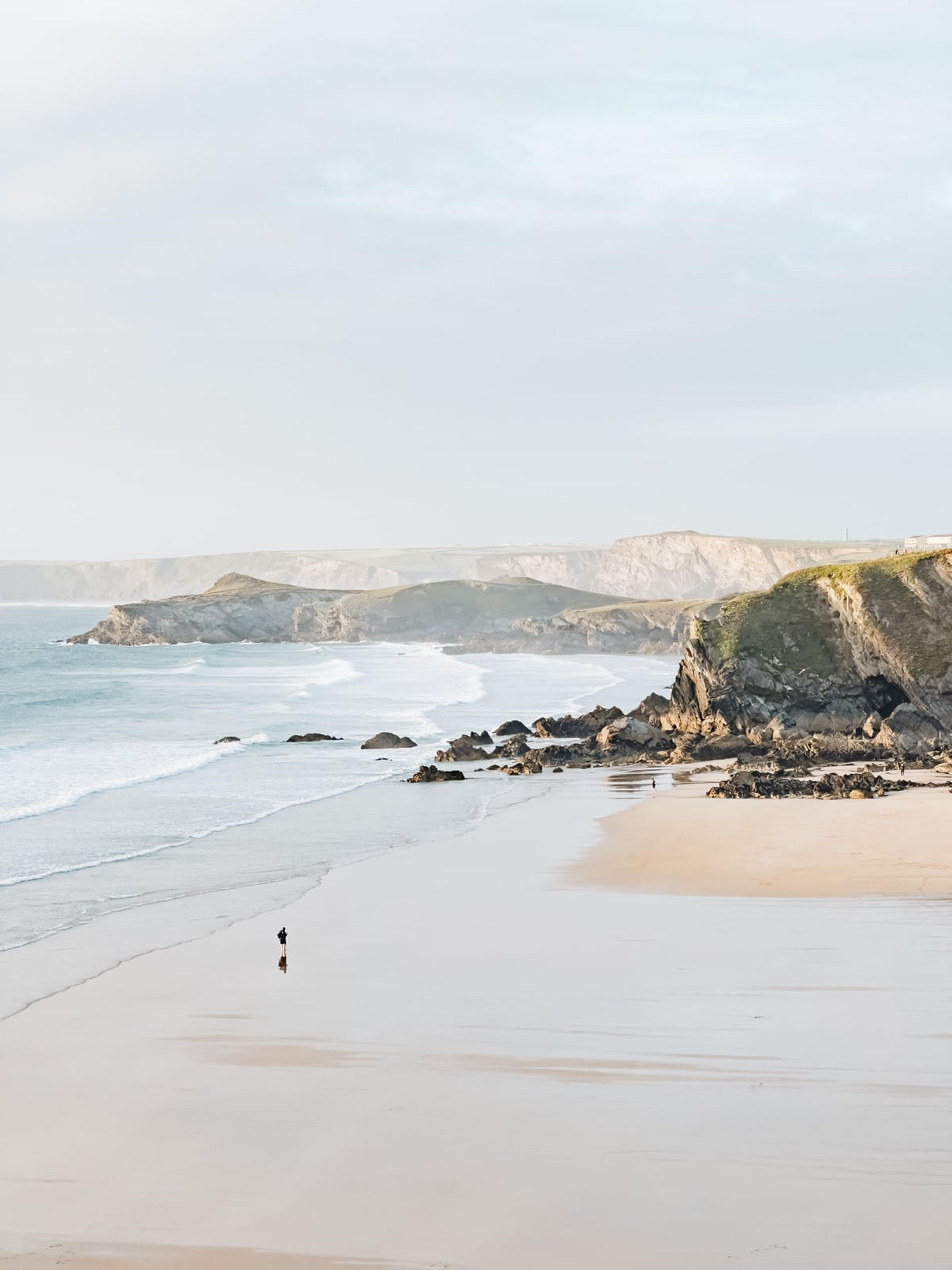 Sandy beach with cliffs and calm surf, a lone figure walking on wet sand in soft morning light