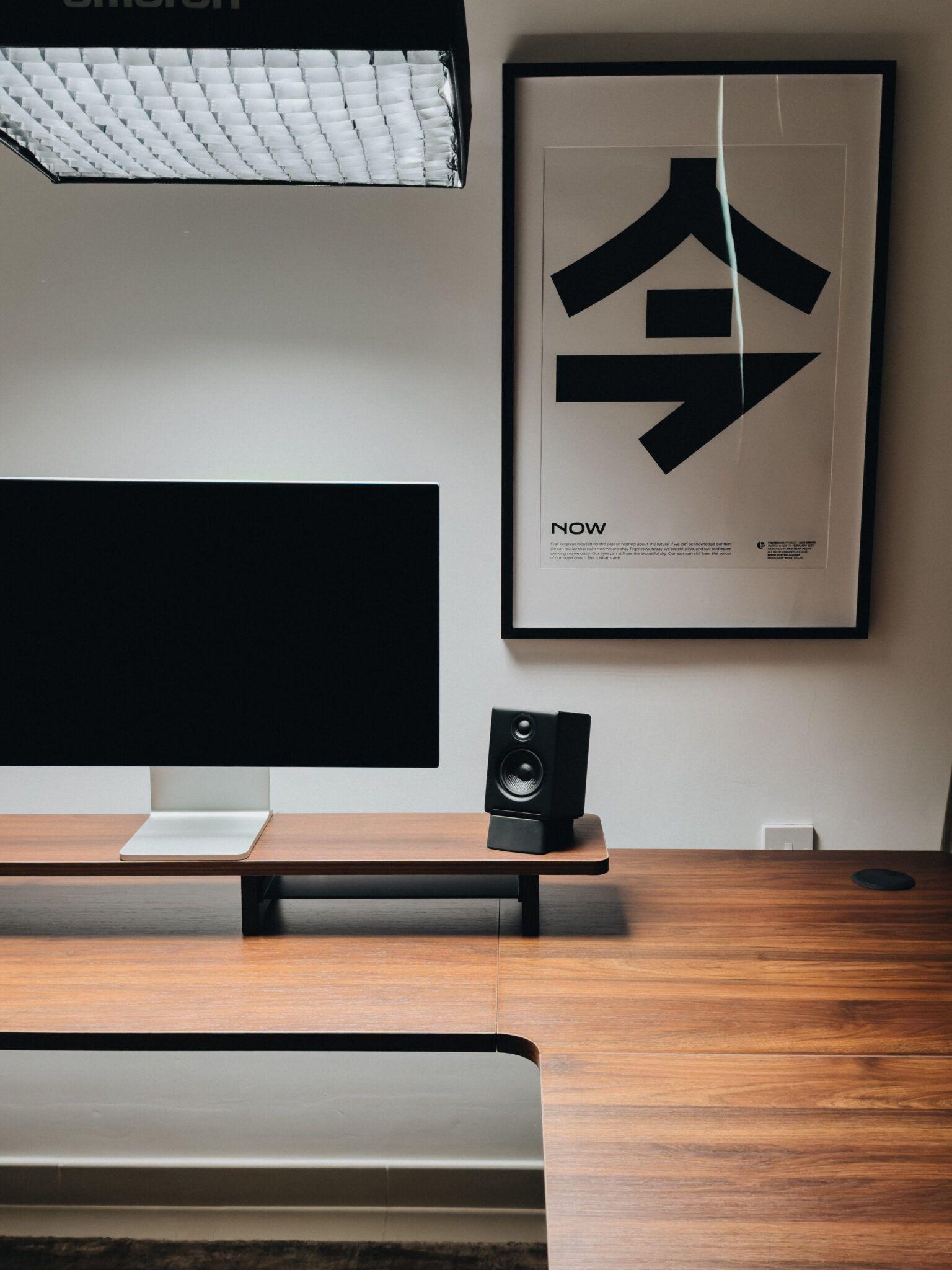 Minimalist desk setup with a monitor stand, small speaker and a graphic 'NOW' poster lit by a softbox overhead