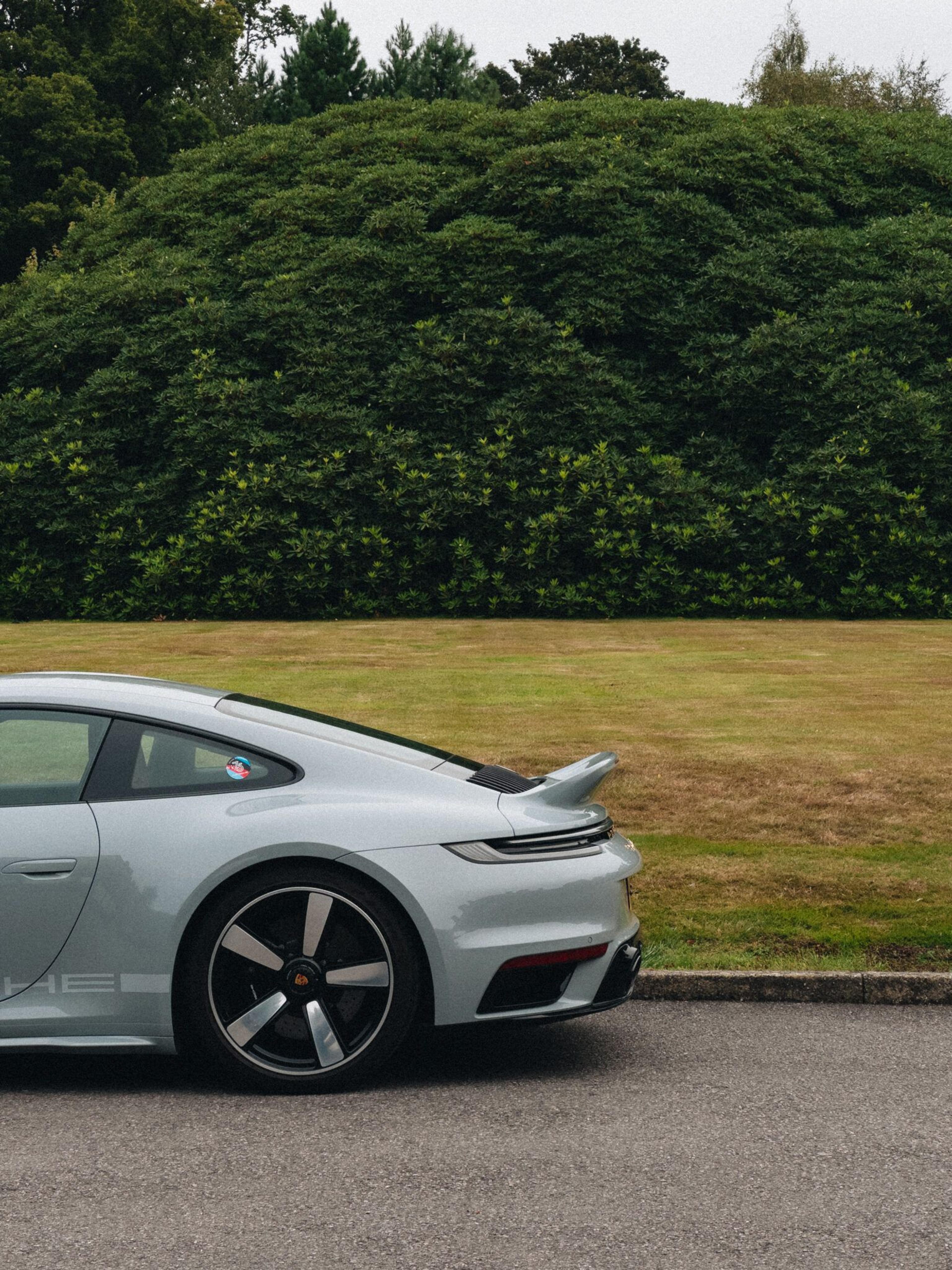 Close rear view of a pale silver sports car with a raised spoiler, parked beside a trimmed hedge and lawn
