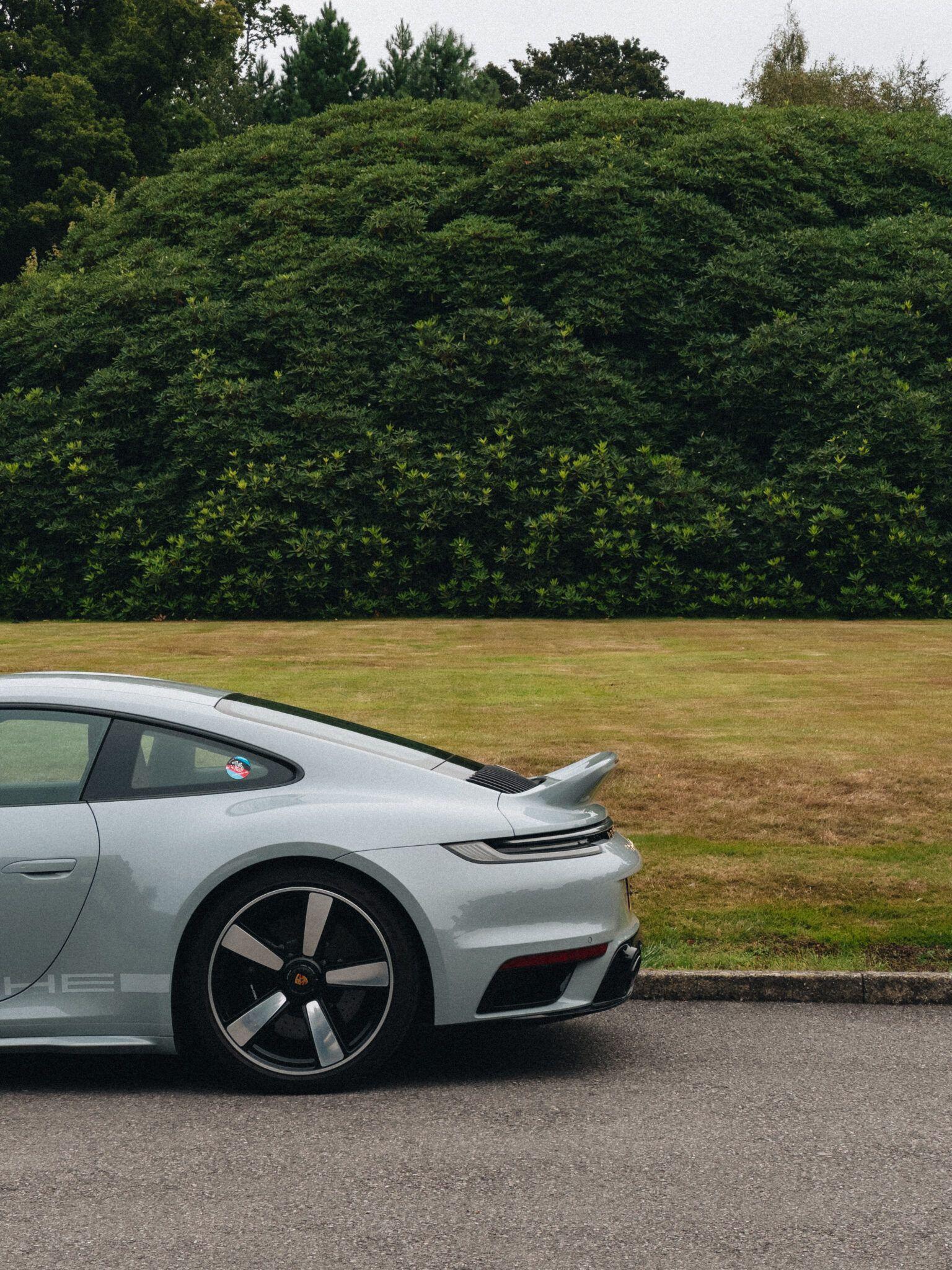 Close rear view of a pale silver sports car with a raised spoiler, parked beside a trimmed hedge and lawn