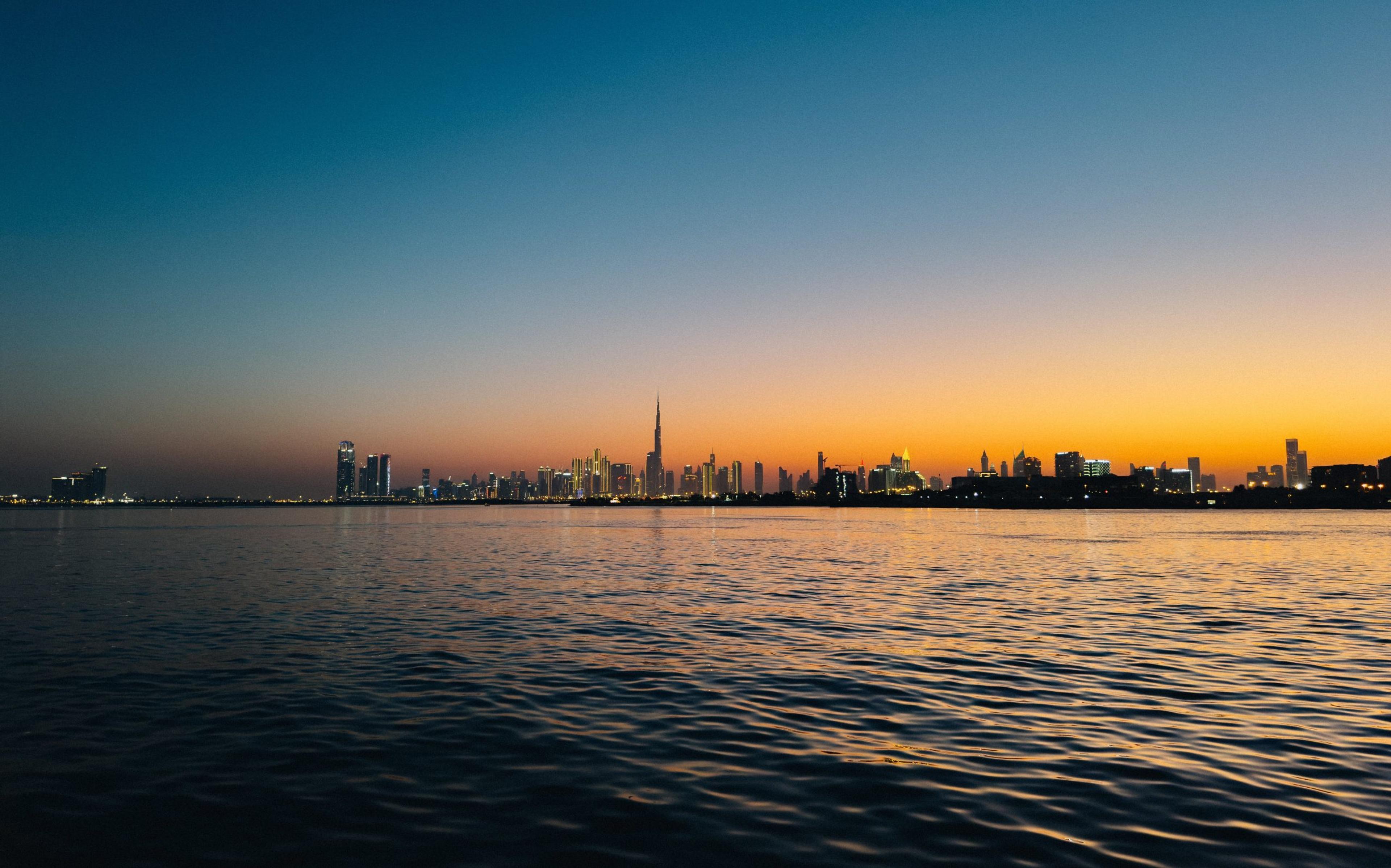 City skyline silhouette with a tall spired tower, glowing orange sunset reflecting on calm water