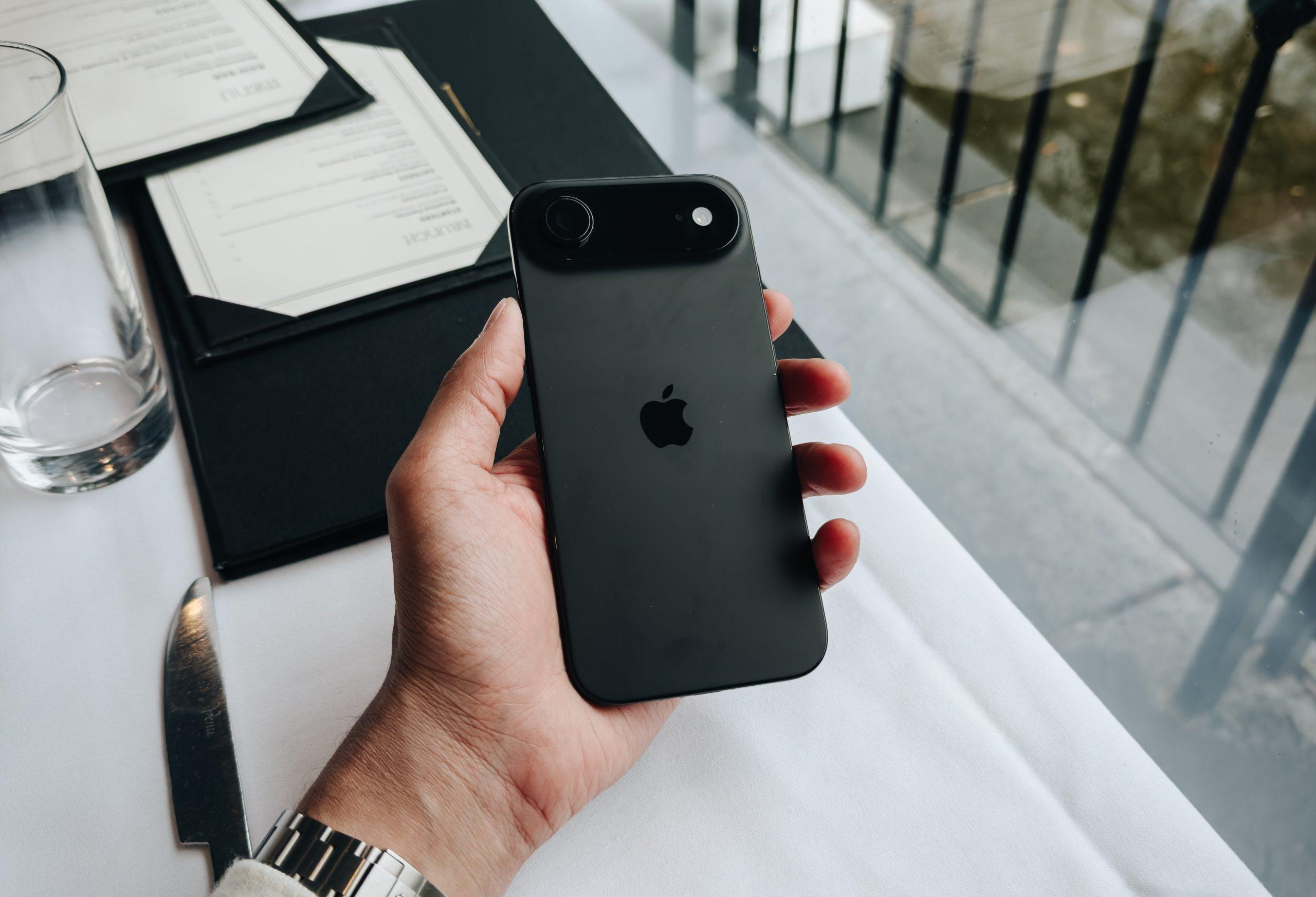 A hand holding a black iPhone, showing its camera lenses, at a restaurant table