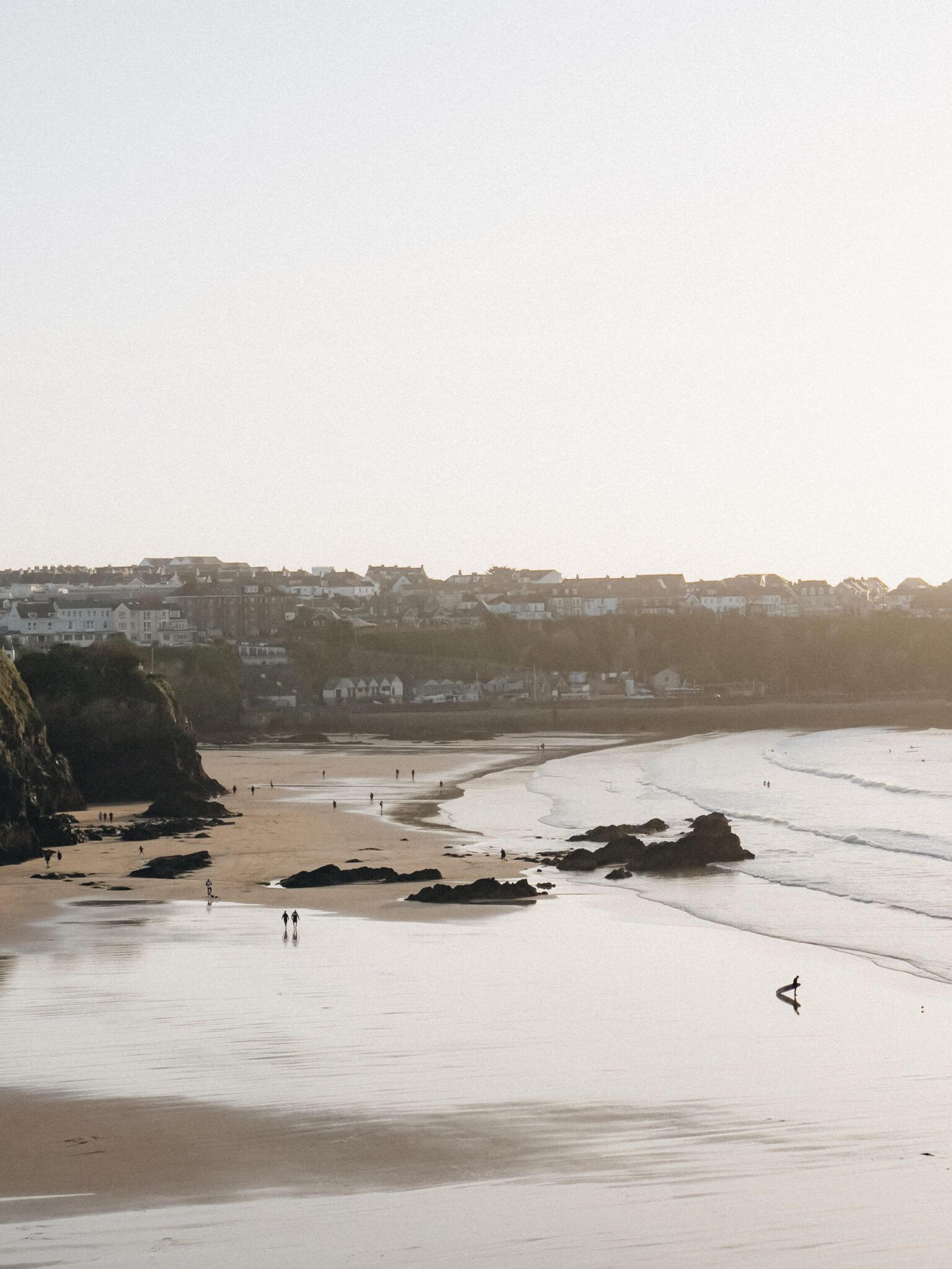 Sandy beach curving past a coastal town at soft golden hour, with a few people and a surfer walking near the shore