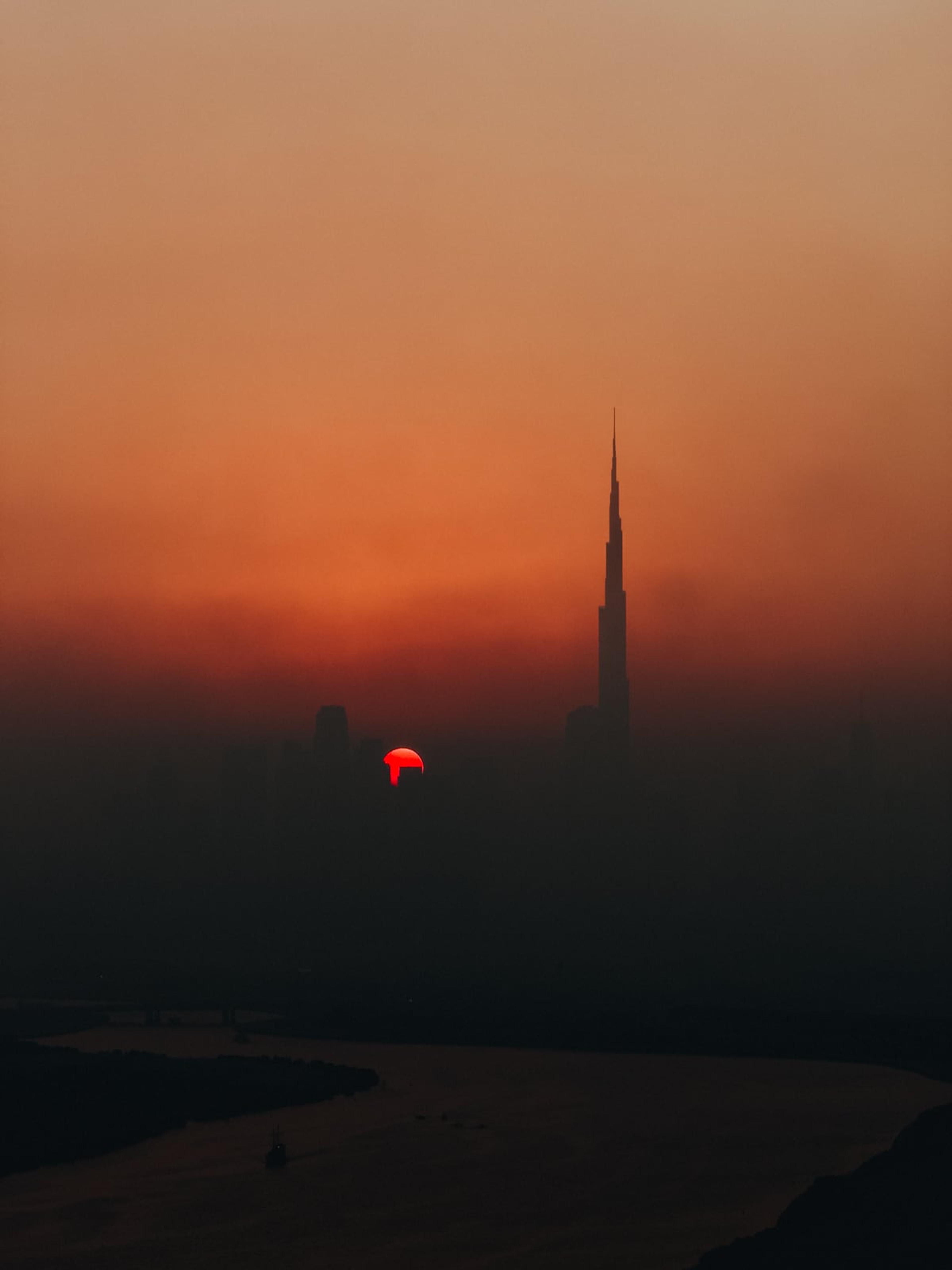 The Burj Khalifa silhouetted against a hazy orange sunset, with a red sun low on the horizon