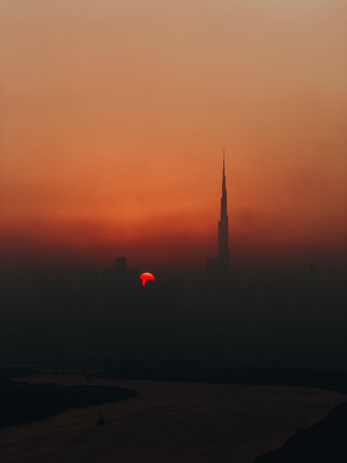 The Burj Khalifa silhouetted against a hazy orange sunset, with a red sun low on the horizon