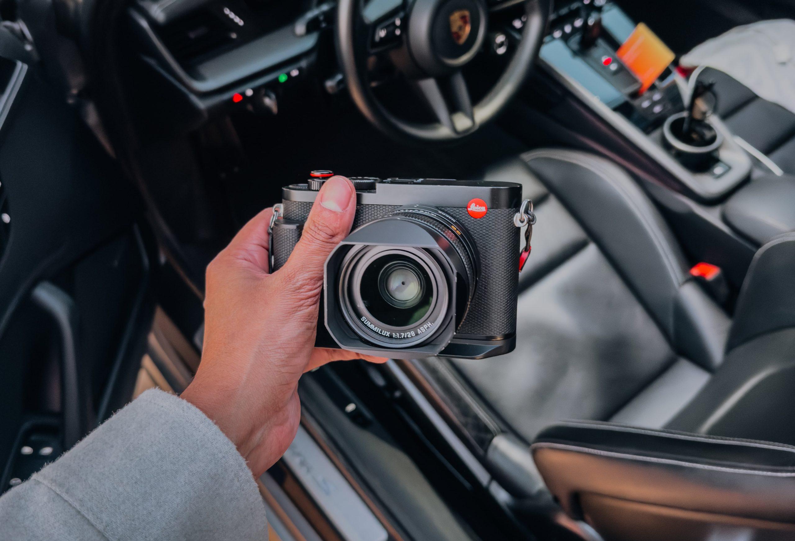 Hand holding a black compact camera inside a car, with the steering wheel and dashboard softly blurred behind it