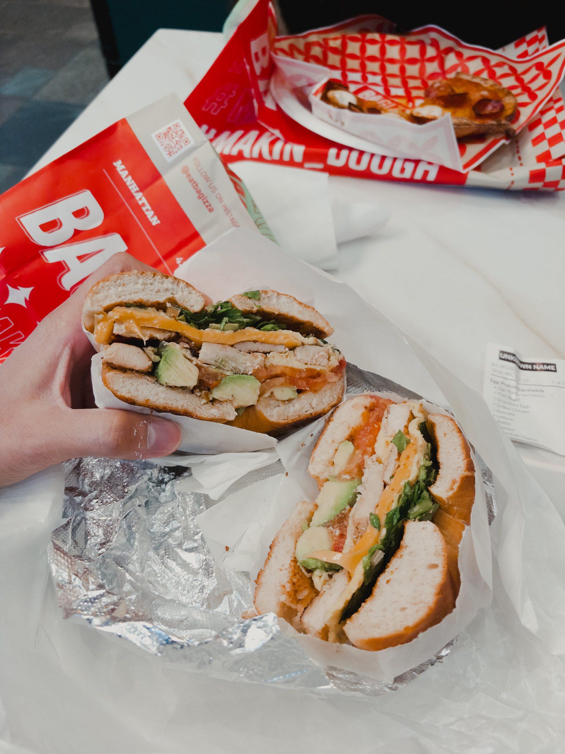 Hand holding half of a stacked deli sandwich with visible fillings, wrapped street food in the background