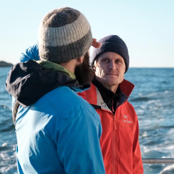 Two people wearing blue and orange coats standing together, talking. Sea in the background. 