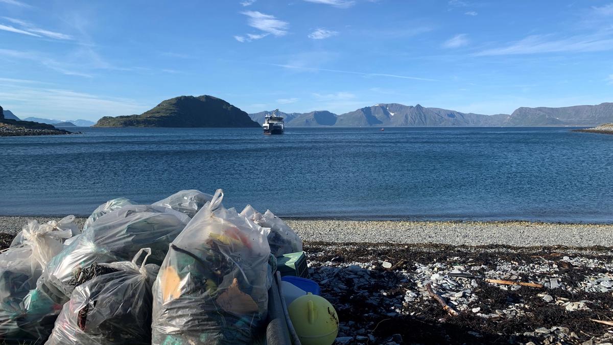  Plastic bags with cleared marine litter on a pebble beach in the foreground, the sea and a large boat in the background
