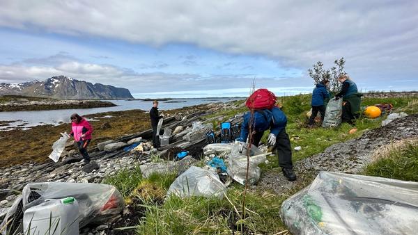 Flere personer rydder forsøpling i søppelsekker i fjæra. Fjell og hav i bakgrunnen