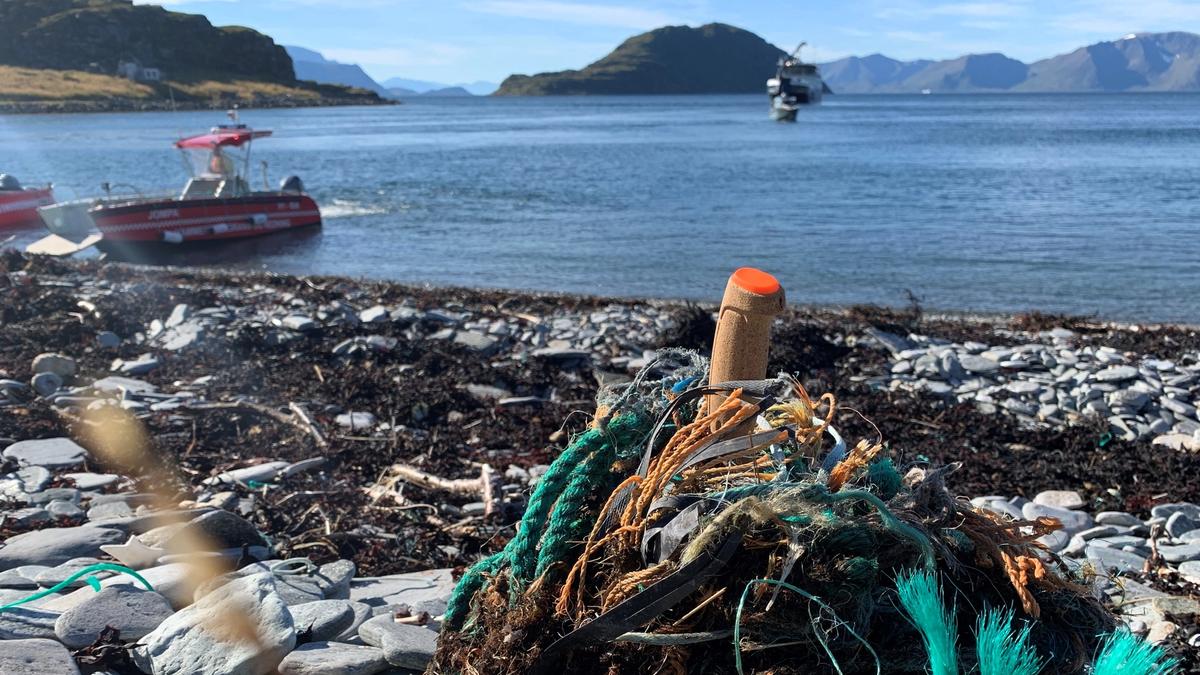 Cluster of ropes, lines and plastic rubbish on a pebble beach with boats and the fjord in the background.