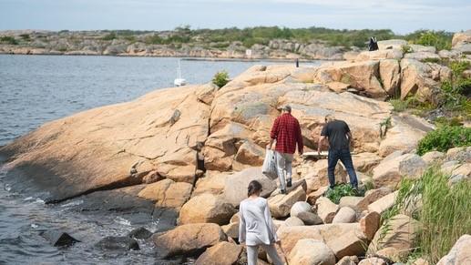 Tre mennesker som rydder søppel ved havet, terrenget er store steiner og svaberg.