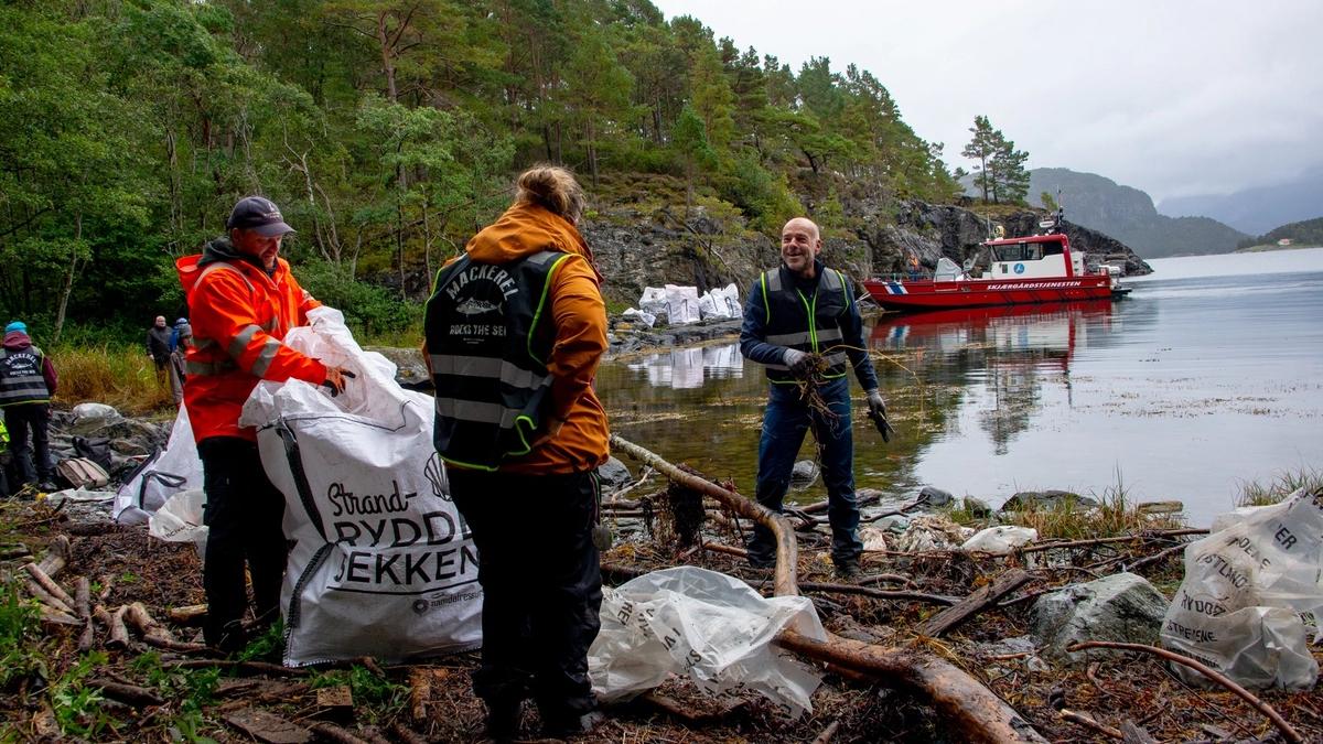 Personer står ved vannkanten til en fjord og rydder søppel. Skjærgårdstjenestens båt i bakgrunnen.