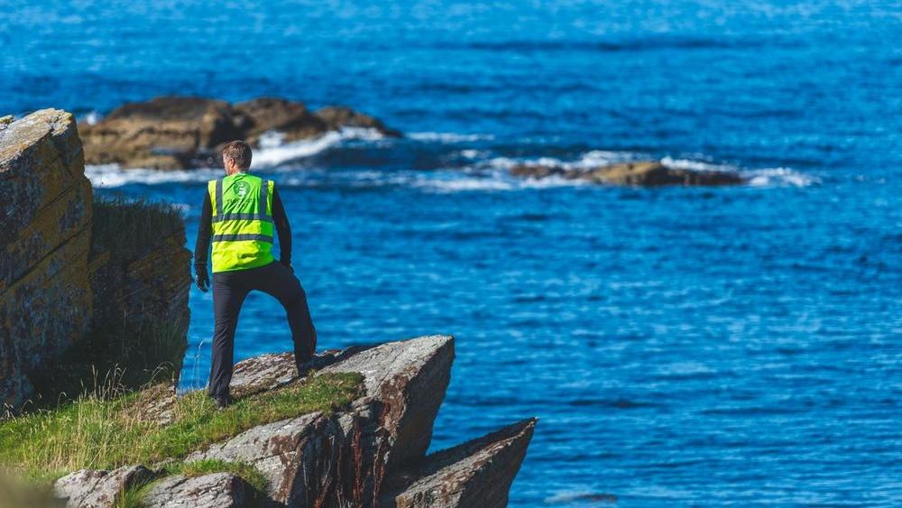 Man in yellow vest is standing on a low cliff facing the sea, his back to the camera