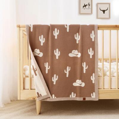 Brown blanket with white cactus and cowboy hat patterns draped over a crib in a nursery.