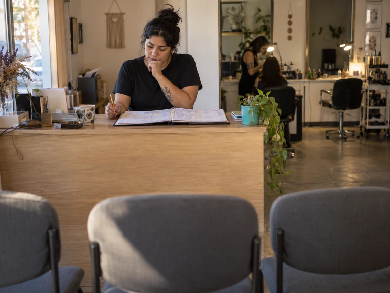 Una recepcionista revisa con un lápiz en la mano la agenda de papel del día, llena de horas anotadas, sobre el mesón de un centro de servicios; al fondo, una de las dos estaciones de trabajo está ocupada y la otra permanece vacía.