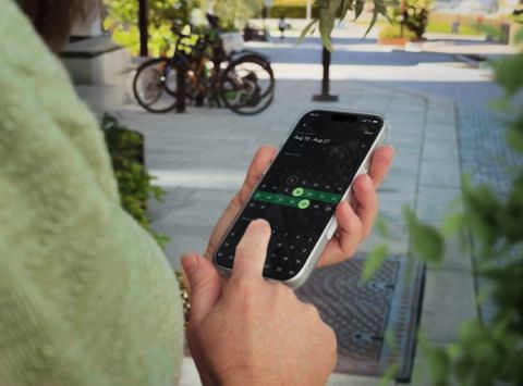 Person in a light green sweater holding a smartphone displaying the Parqio app's date-range calendar picker, with bicycles and a sunny urban footpath visible in the background.