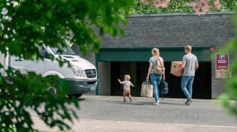 Two adults carrying bags and a box walk with a small child toward an underground parking garage entrance beside a parked white van, framed by green foliage.