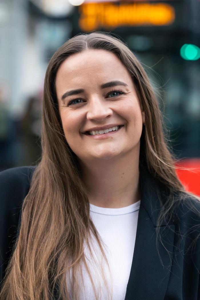 Smiling person with long brown hair and a black blazer stands outdoors in an urban setting with blurred lights and buildings in the background.